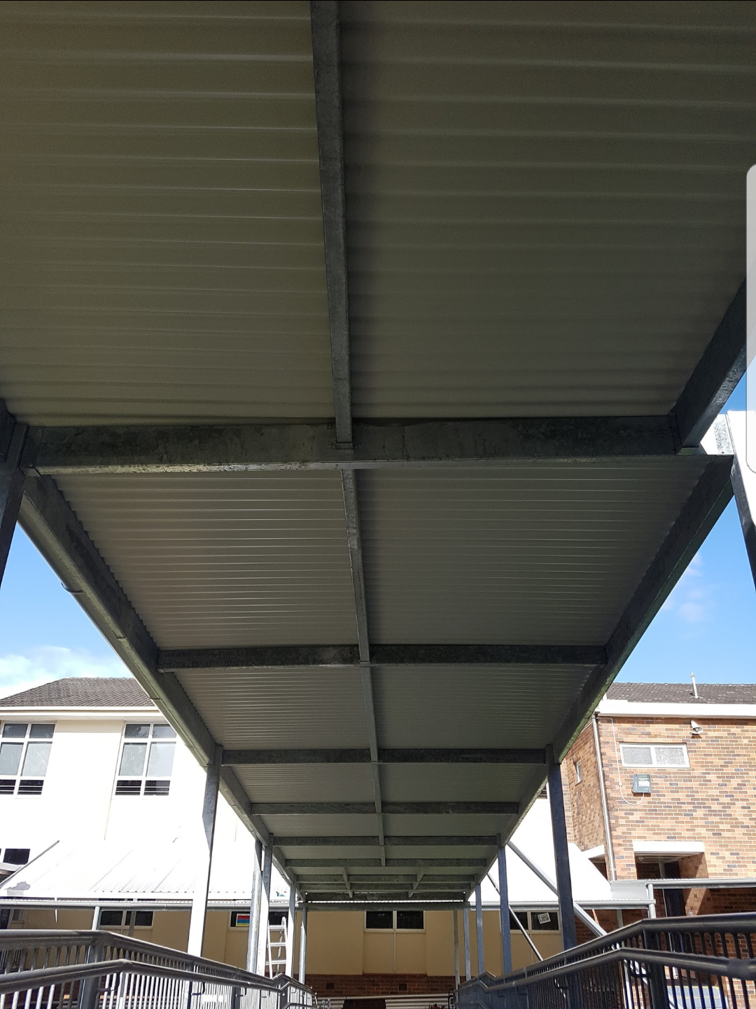 View underneath a metal canopy with a corrugated roof, supported by vertical and cross beams, outside a building with brick and painted walls, and a clear blue sky in the background.
