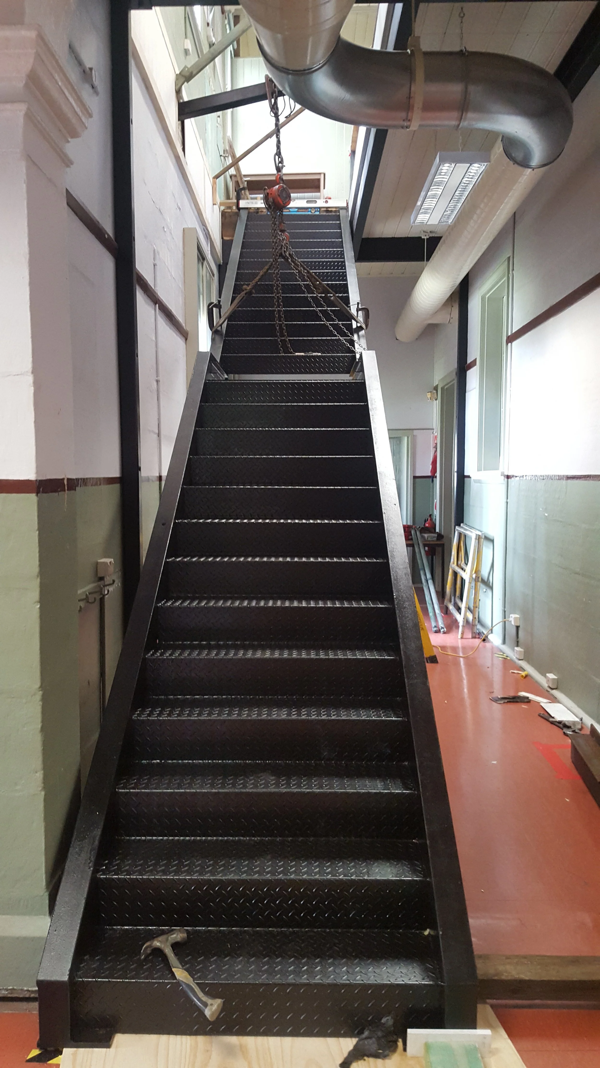 Black metal staircase with a hand railing, ascending in an industrial interior space. construction tools and equipment are on the floor and stairs, including a hammer and tape measure. Pipes and window frames are visible on the walls.