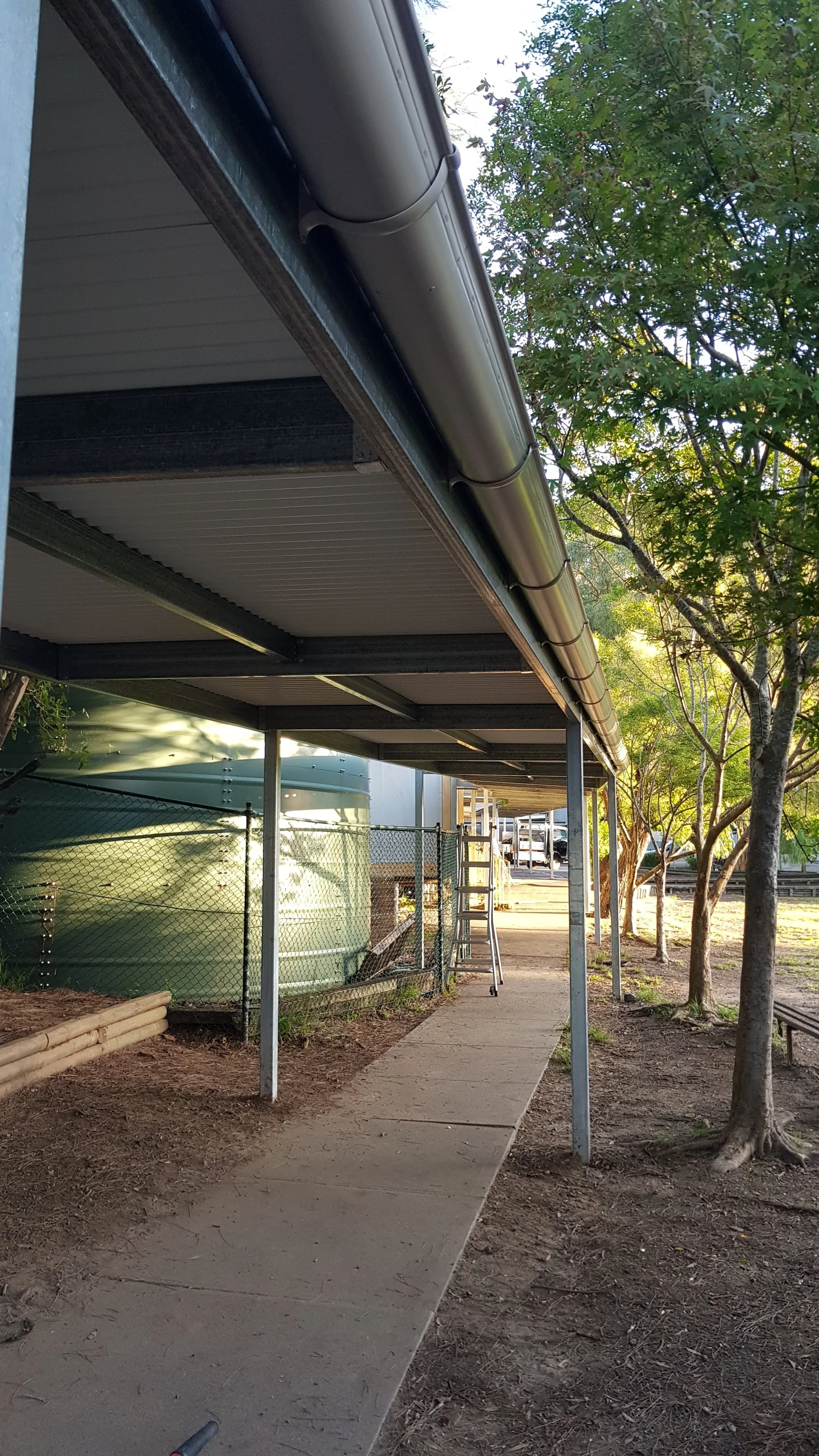 Side view of a modern building with a metal roof and gutter system, supported by metal posts, with a sidewalk and trees outside.