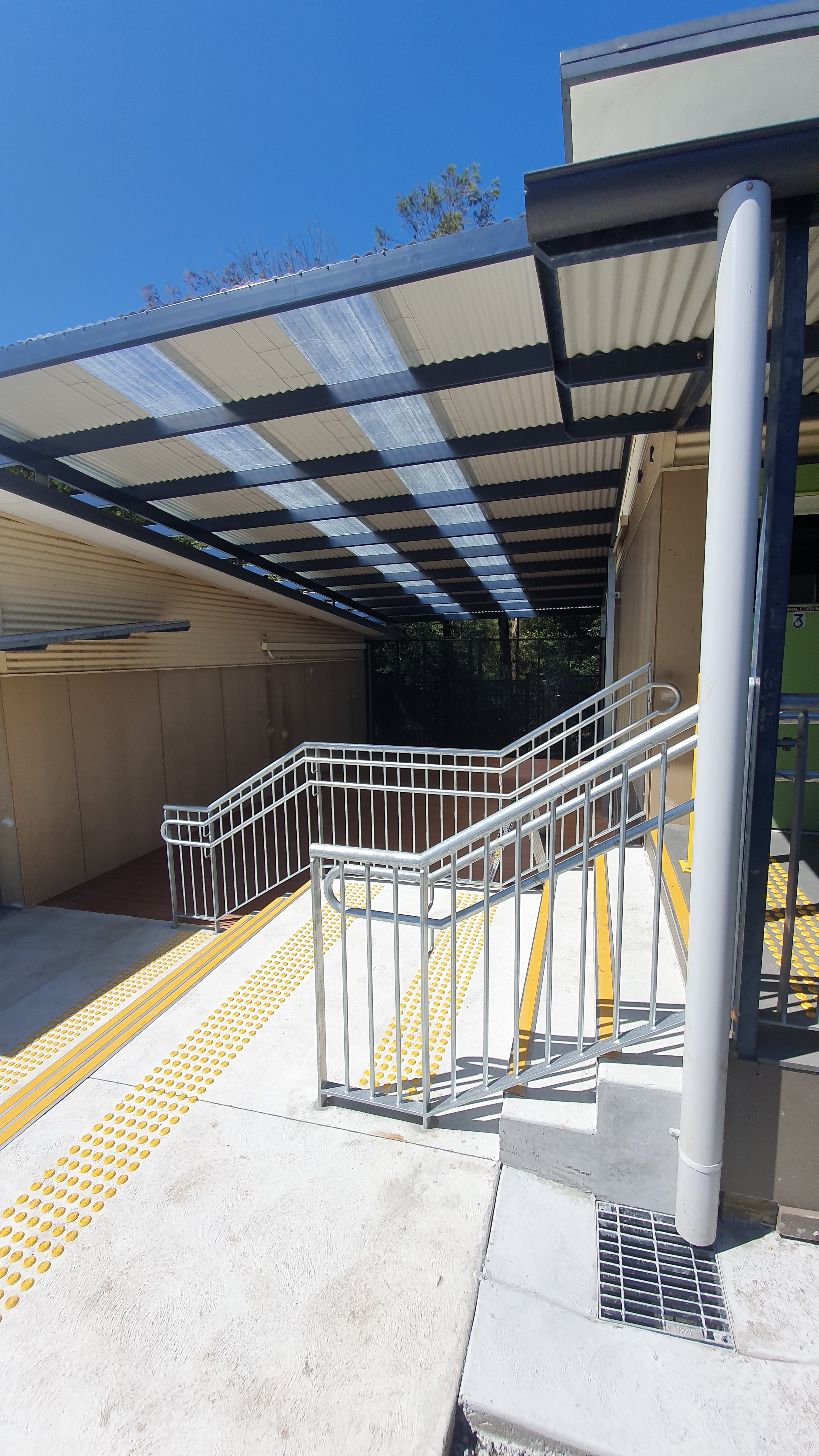 A modern outdoor staircase with yellow tactile paving, metal railings, and a covered entryway, leading to a building entrance.