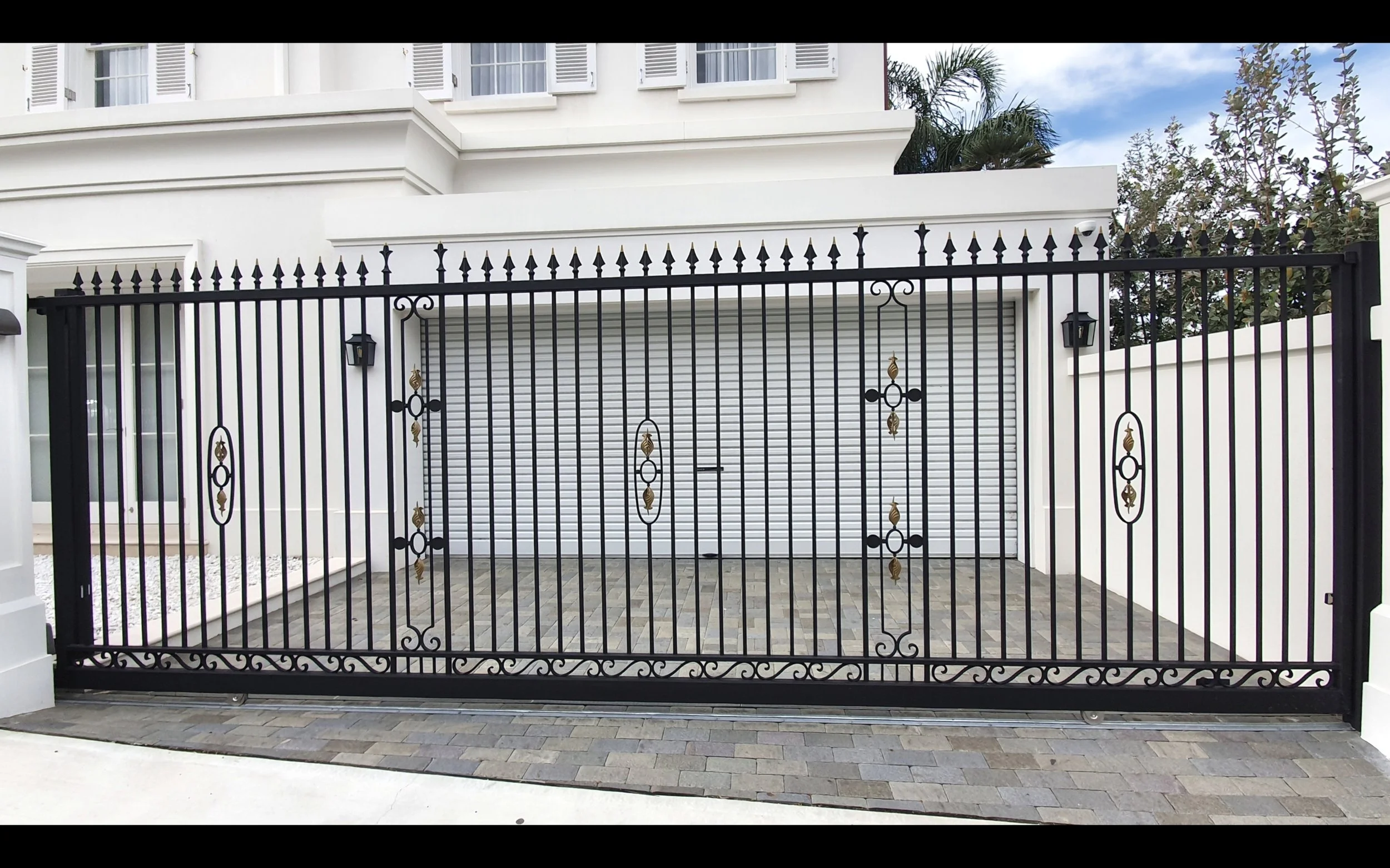 View of the front gate of a white house with a garage door behind it, black decorative iron fence with gold accents, and two wall-mounted lantern lights on either side of the gate.