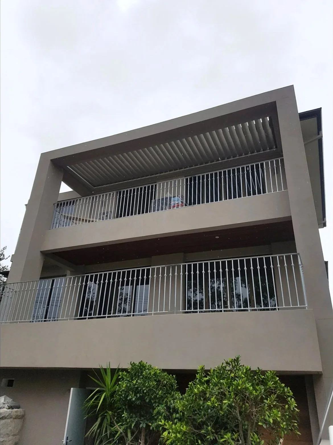 Modern multi-story building with balconies and metal railings, with greenery in the foreground and an overcast sky.