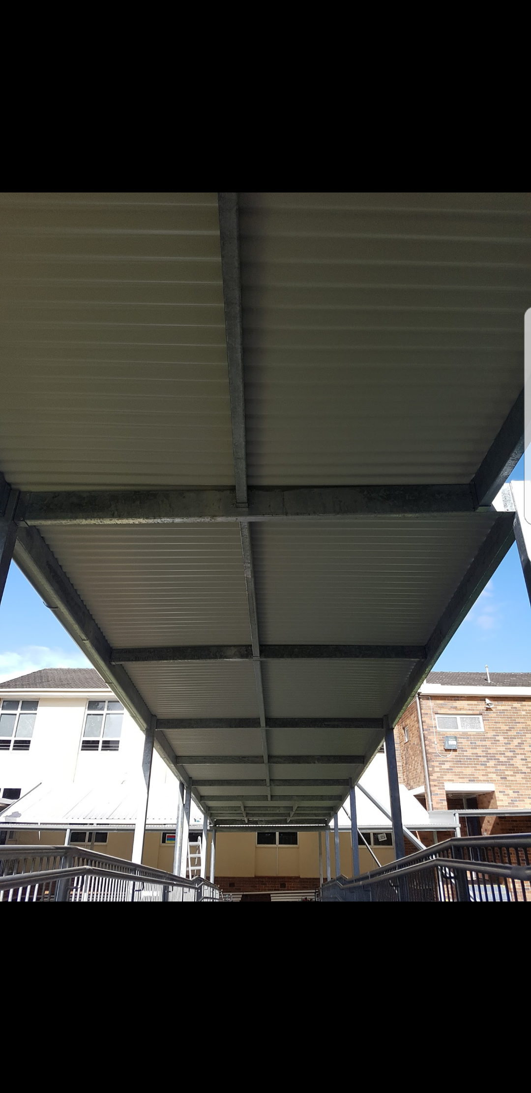 View looking up at the underside of a metal roof structure supported by metal framework in an outdoor area, with buildings visible in the background under a blue sky.