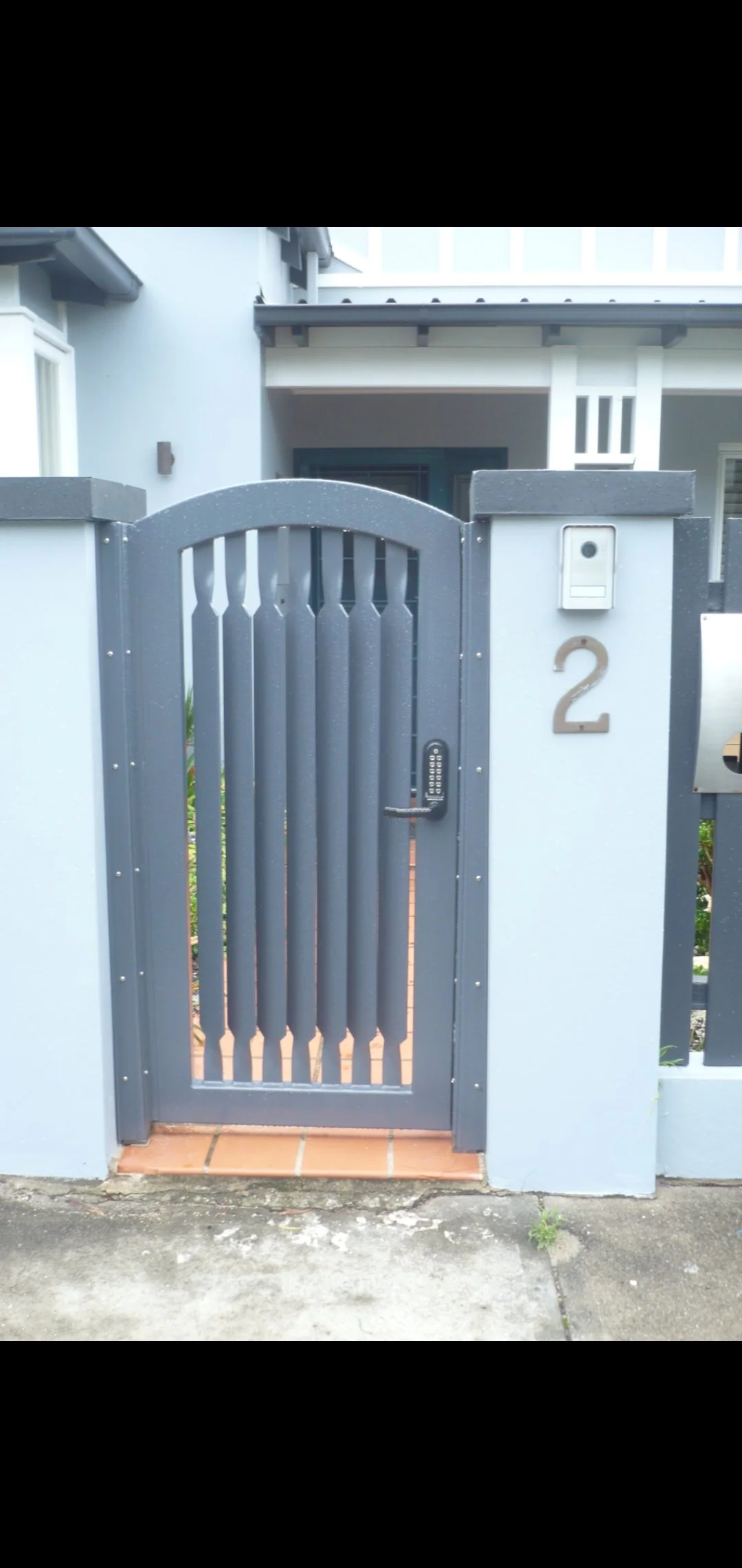 Gray metal gate with digital lock and the number 2 on a white wall, with a house in the background.