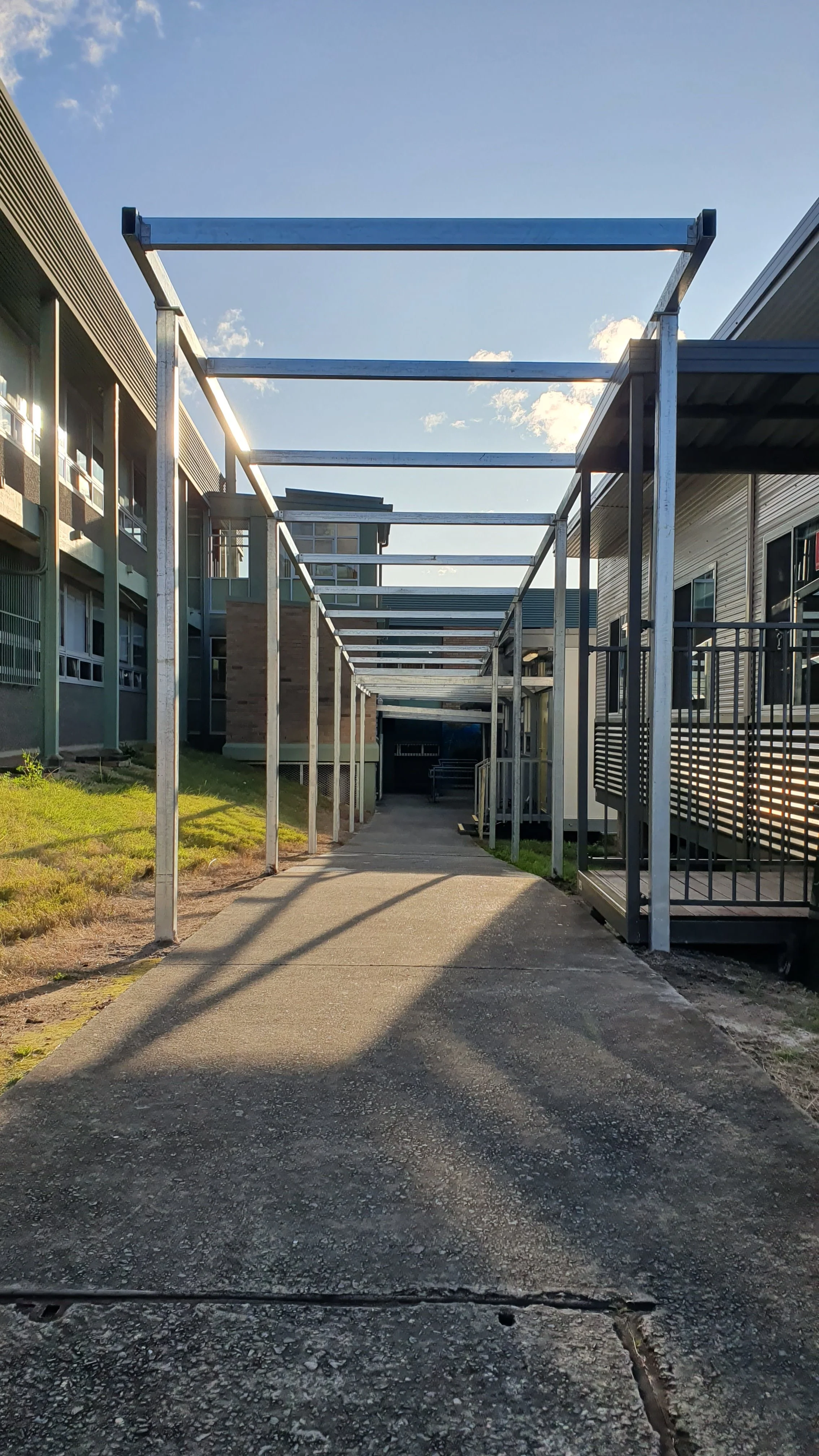 A concrete pathway leading through an outdoor walkway with metal frame supports for a future pergola or canopy, flanked by modern apartment buildings on either side, with a clear blue sky overhead.
