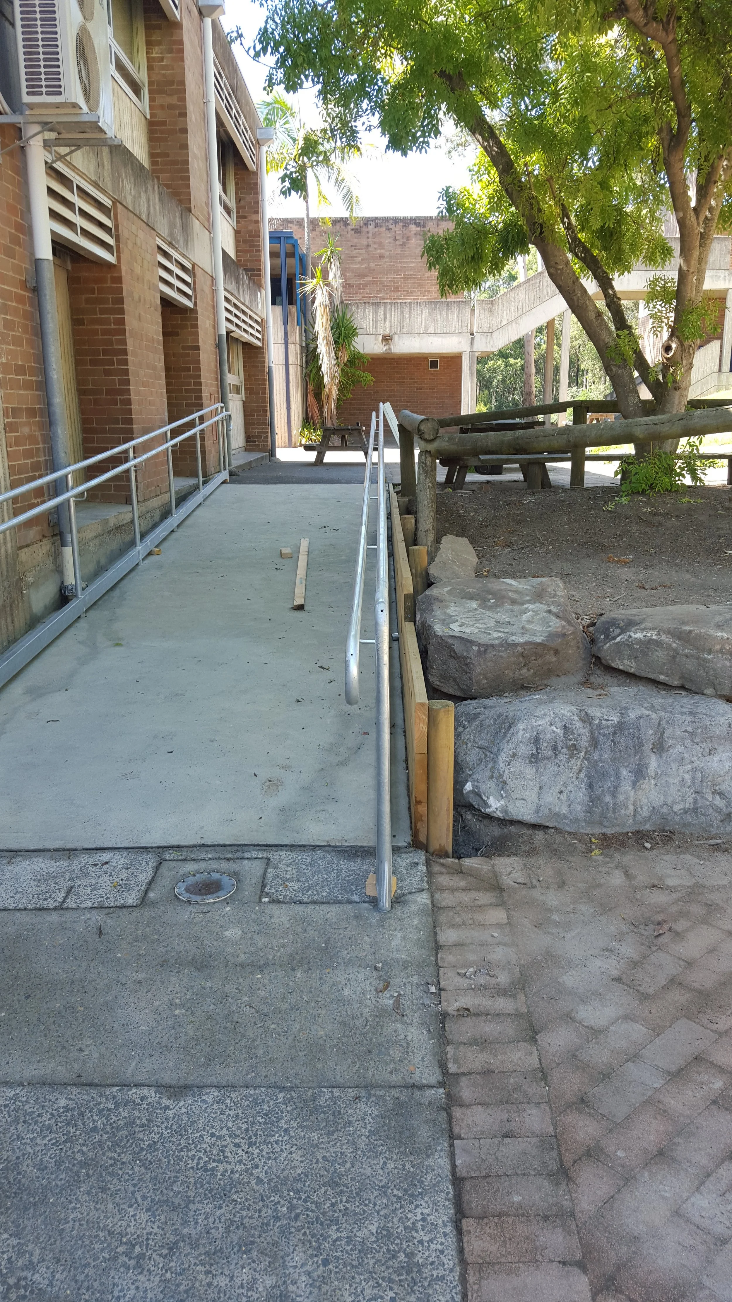 Concrete ramp with metal handrails leading to a building entrance, next to a dirt area with large rocks and a wooden fence, and a tree with green leaves providing shade.