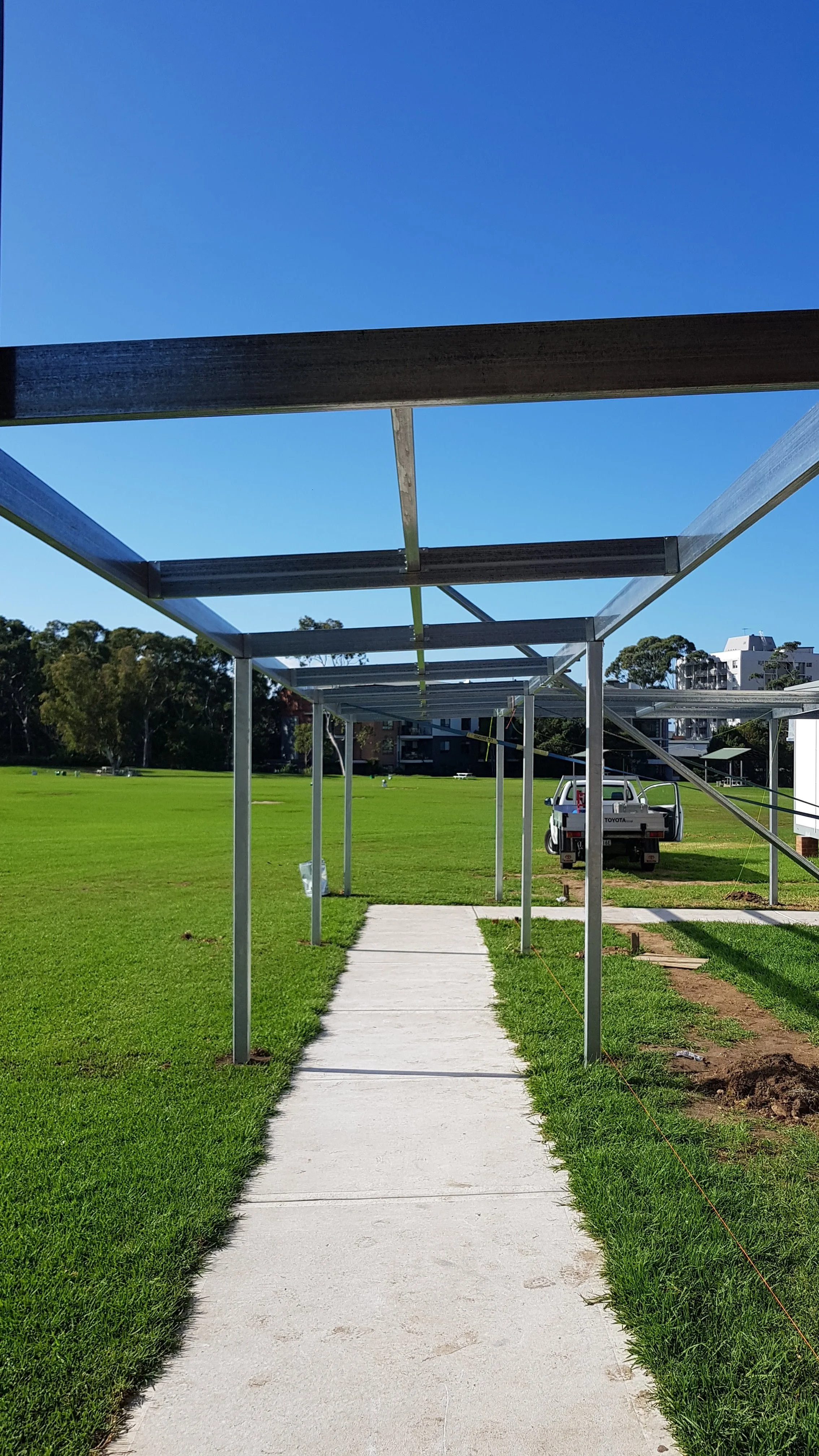 A construction site with a new walkway being built, metal framework for a canopy over the sidewalk, a small white truck, and a grassy field with trees in the background.