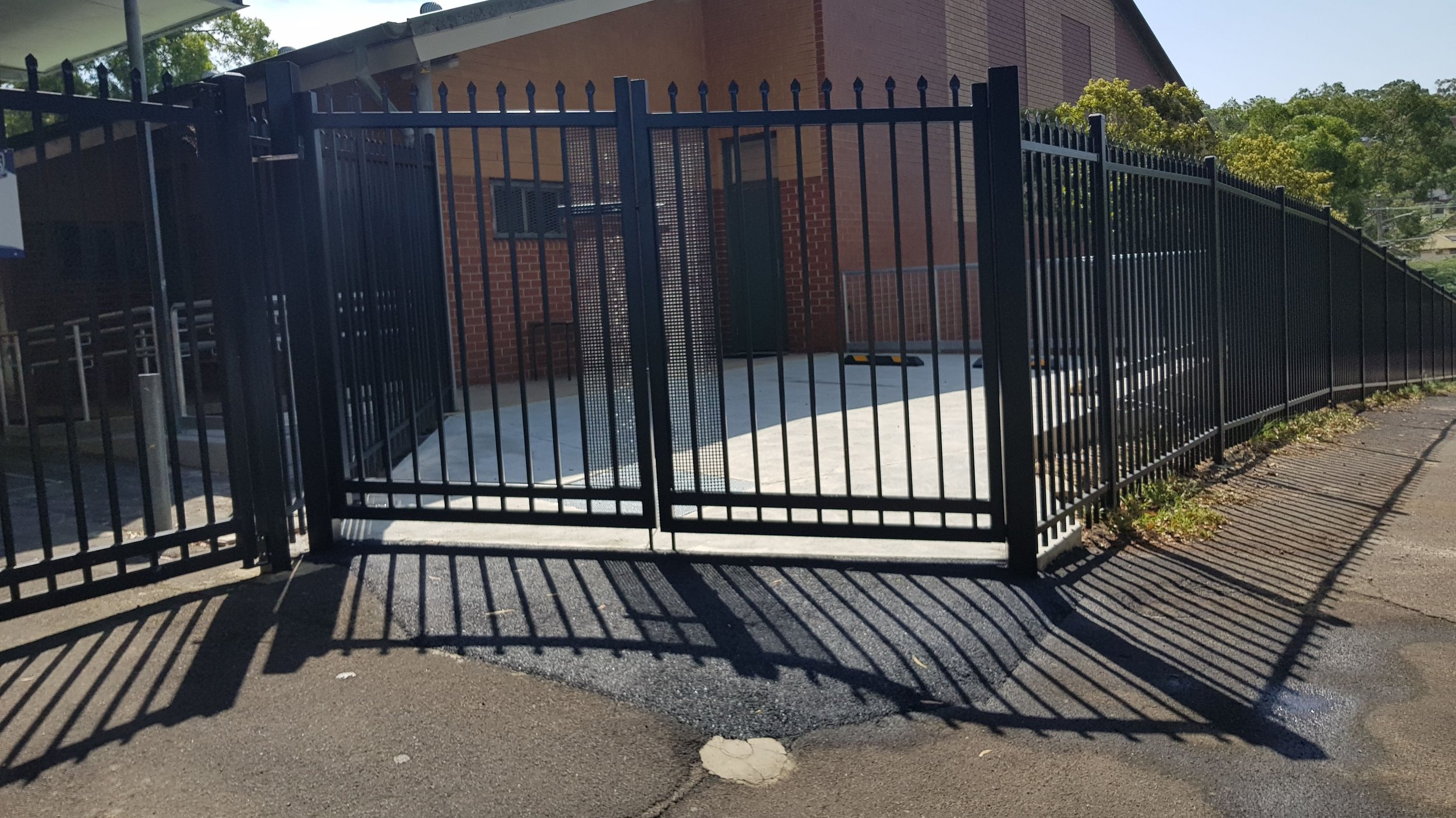 A black metal fence with a sliding gate encloses a parking area near a brick building with a sloped roof. Trees and bushes are visible in the background. Shadows of the fence are cast on the street pavement.