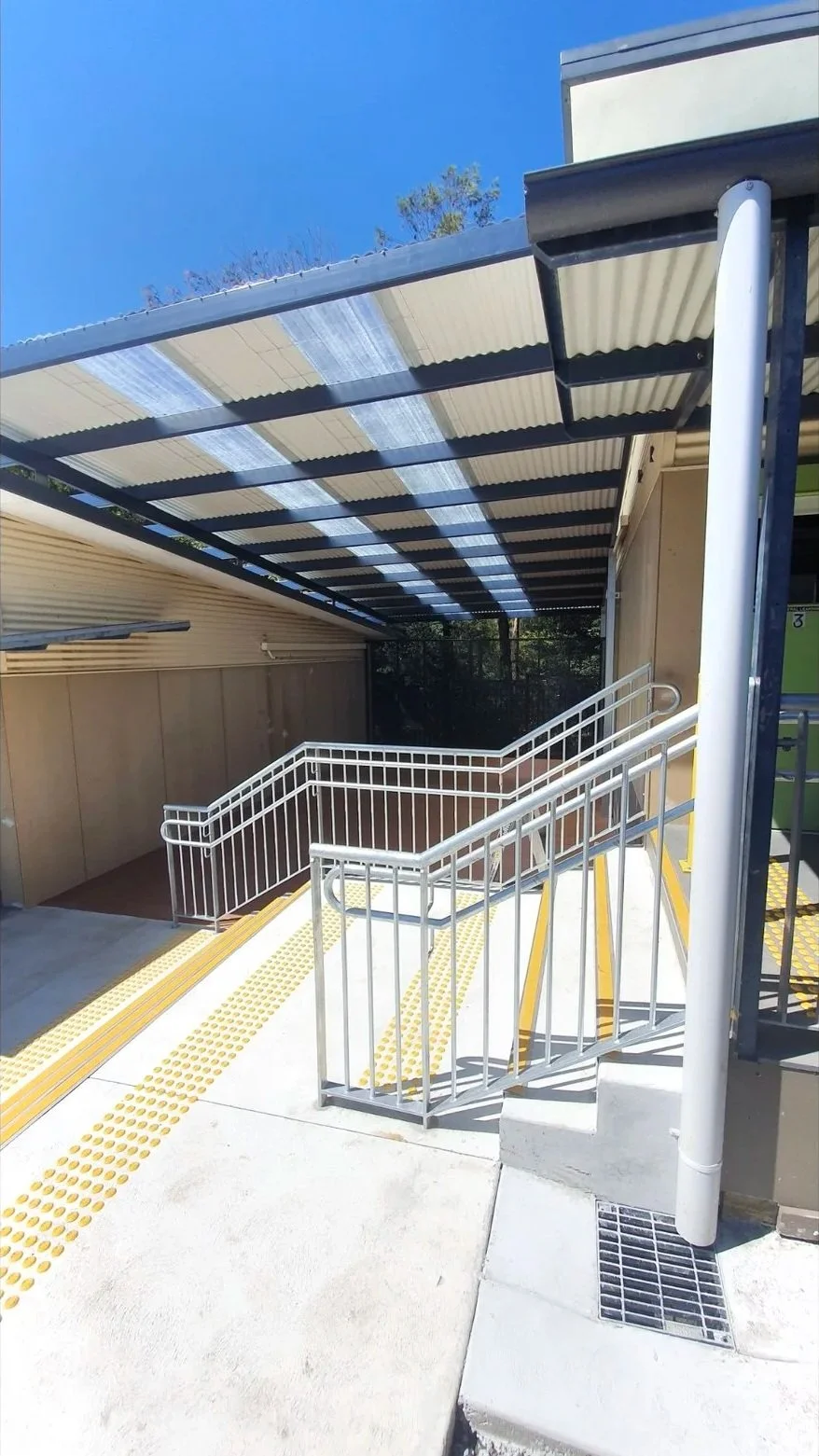 Exterior view of a building with a ramp, handrails, tactile paving, and a partially covered walkway under a blue sky.