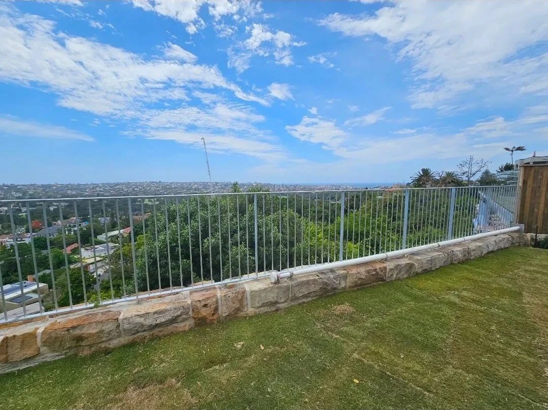View from a grassy yard with a white metal railing, overlooking a suburban landscape with trees and houses, under a partly cloudy blue sky.
