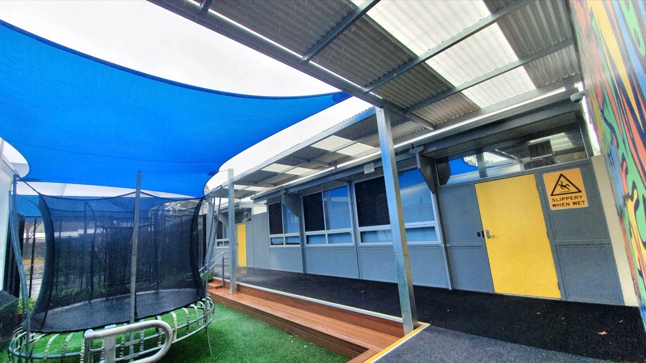 Outdoor area with a blue shade sail canopy, trampoline with safety net, yellow door, and colorful wall art, under a covered patio with a sign that says 'slippery when wet.'