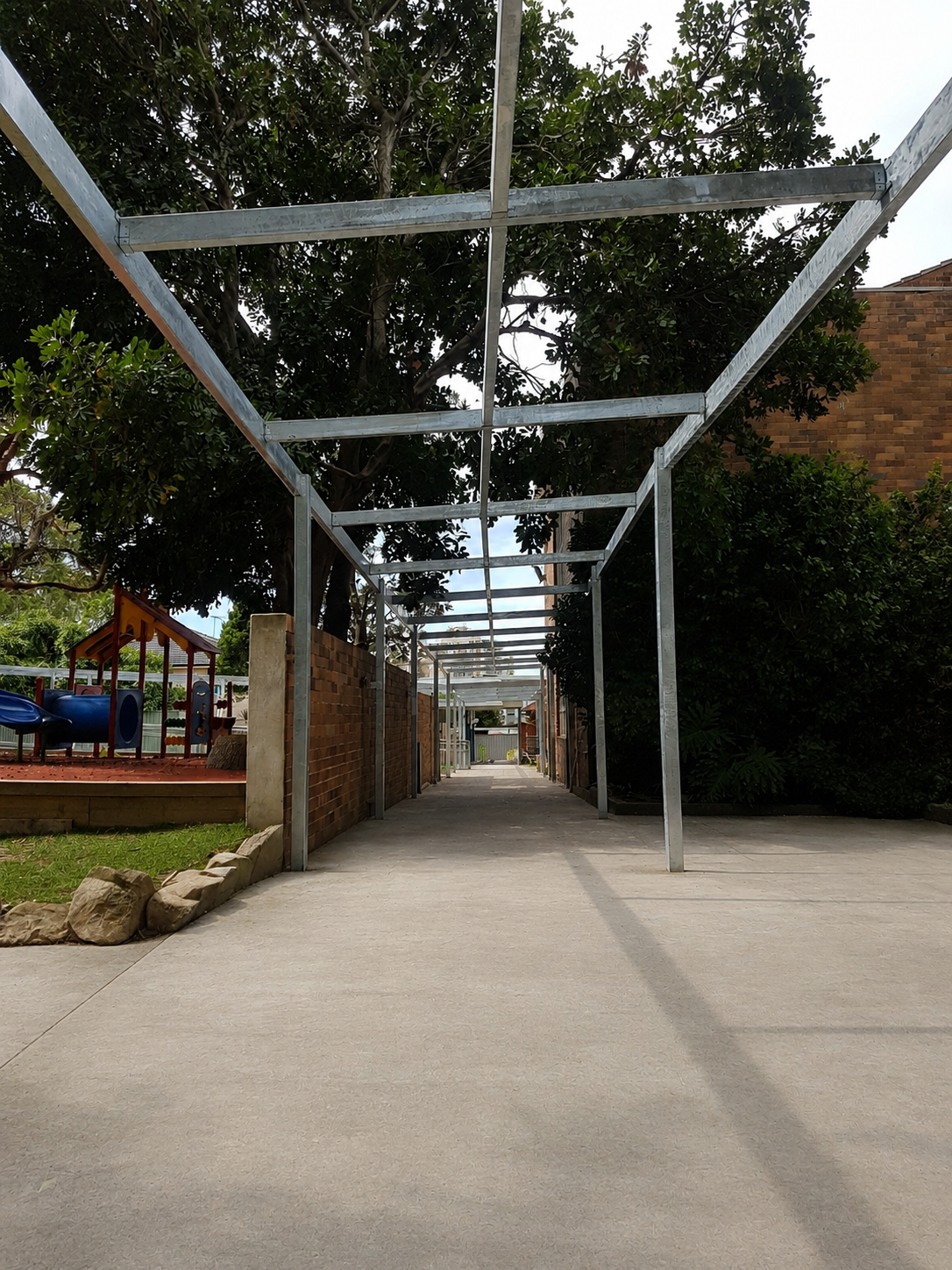 Metal framework structure over a concrete pathway in a backyard, with trees and a brick wall on either side.