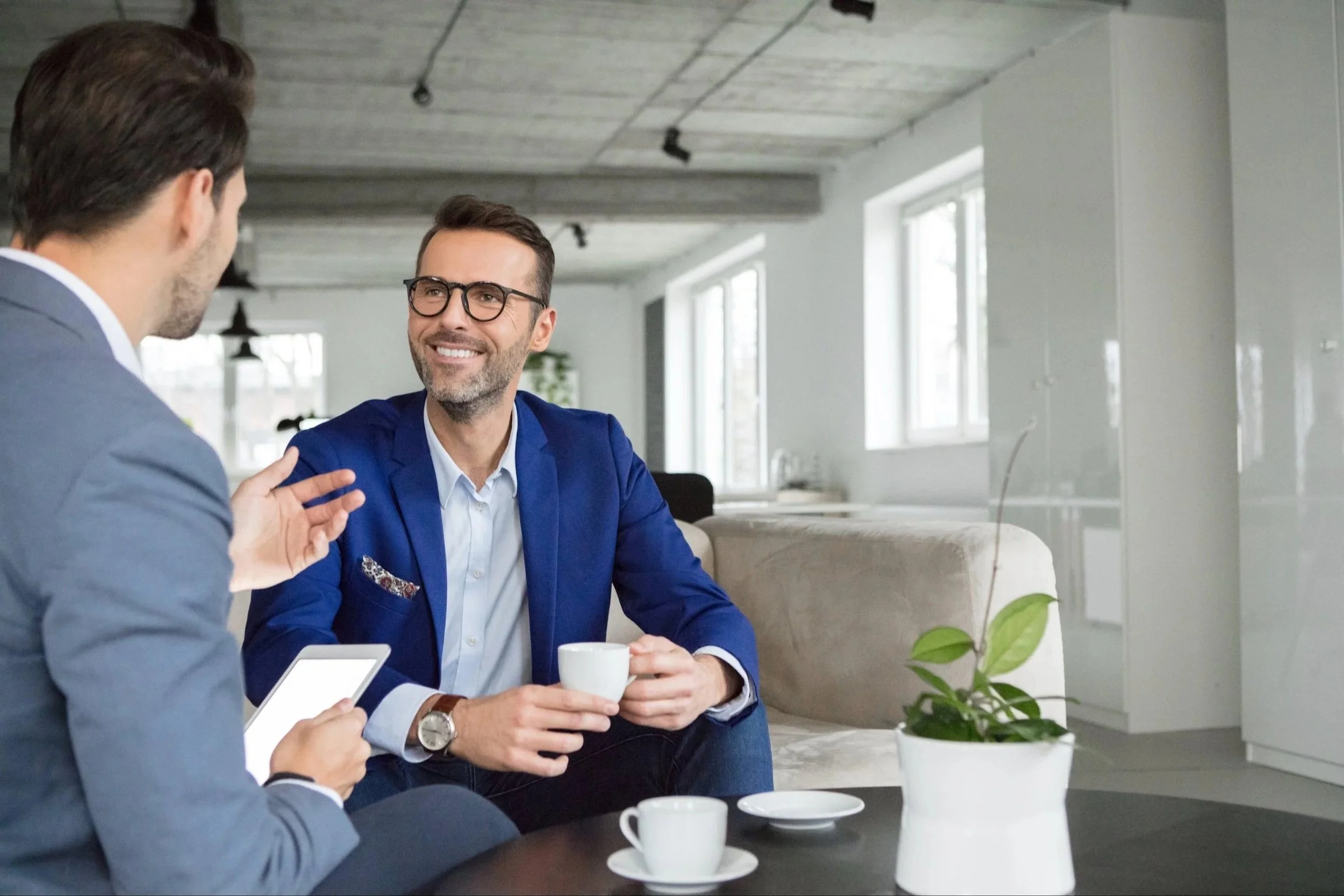 Two men in business attire engaged in conversation, one with glasses and a beard, holding a cup, in a modern office setting with large windows and minimalist decor.