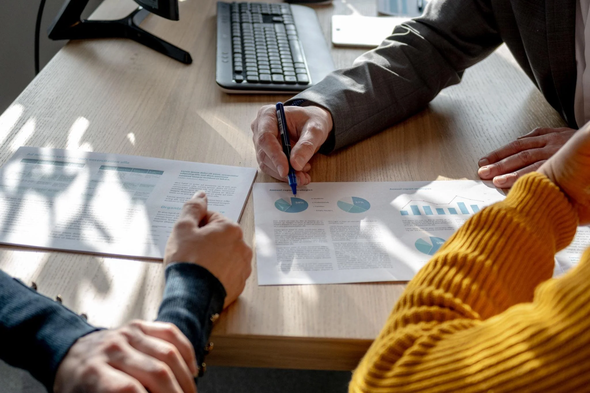 Two people sitting at a wooden table discussing documents with charts and graphs, one person wearing a black jacket and the other in a yellow sweater, sunlight casting shadows on the papers.