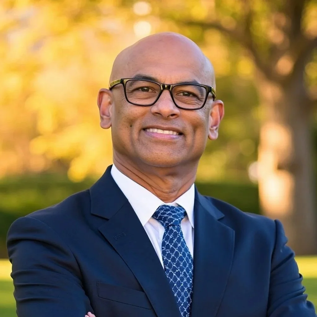 A man in a navy suit and glasses smiling outdoors with trees and warm sunset lighting in the background.