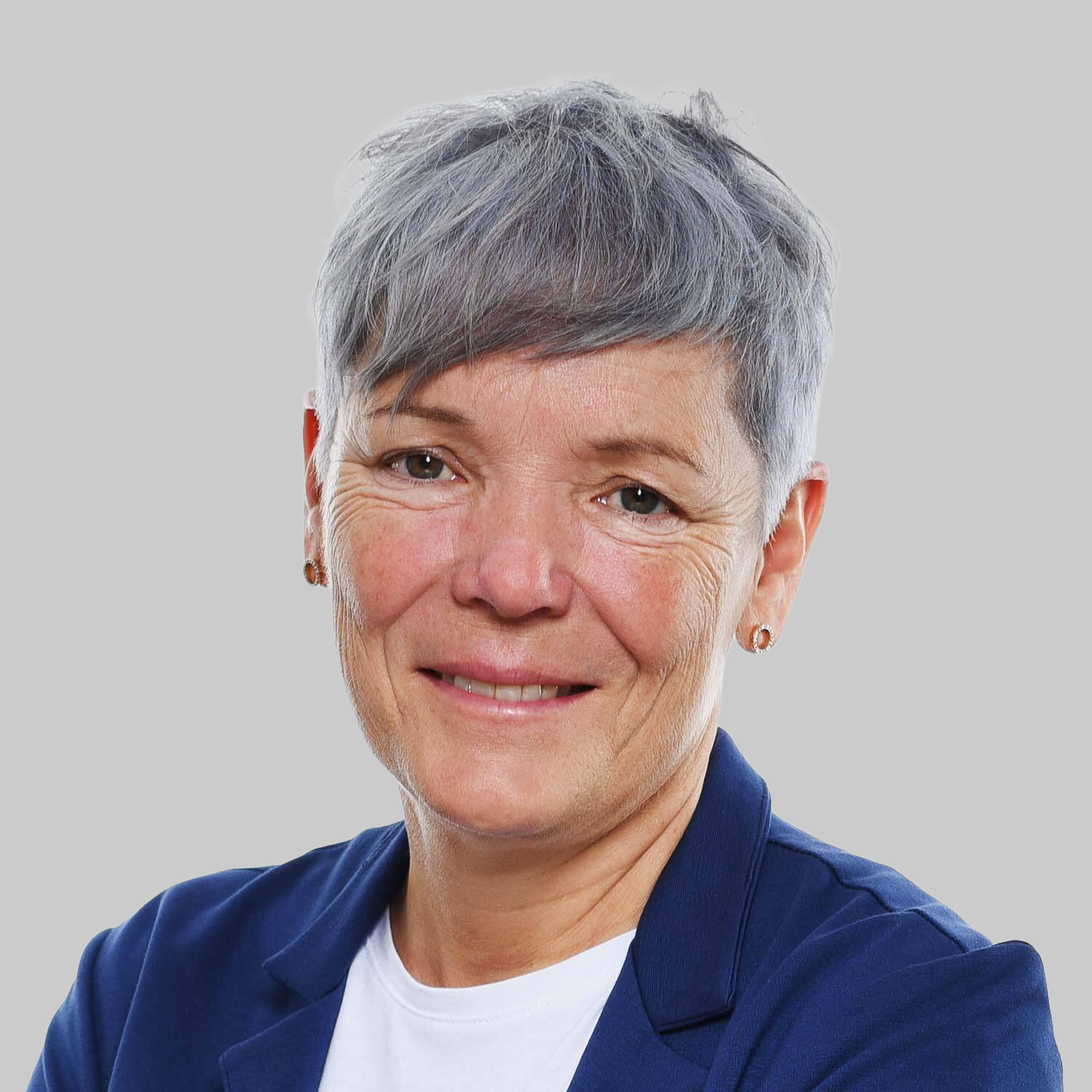 Nahaufnahme eines lächelnden älteren Frau mit kurzen grauen Haaren, die eine dunkelblaue Jacke trägt, vor hellem Hintergrund.
