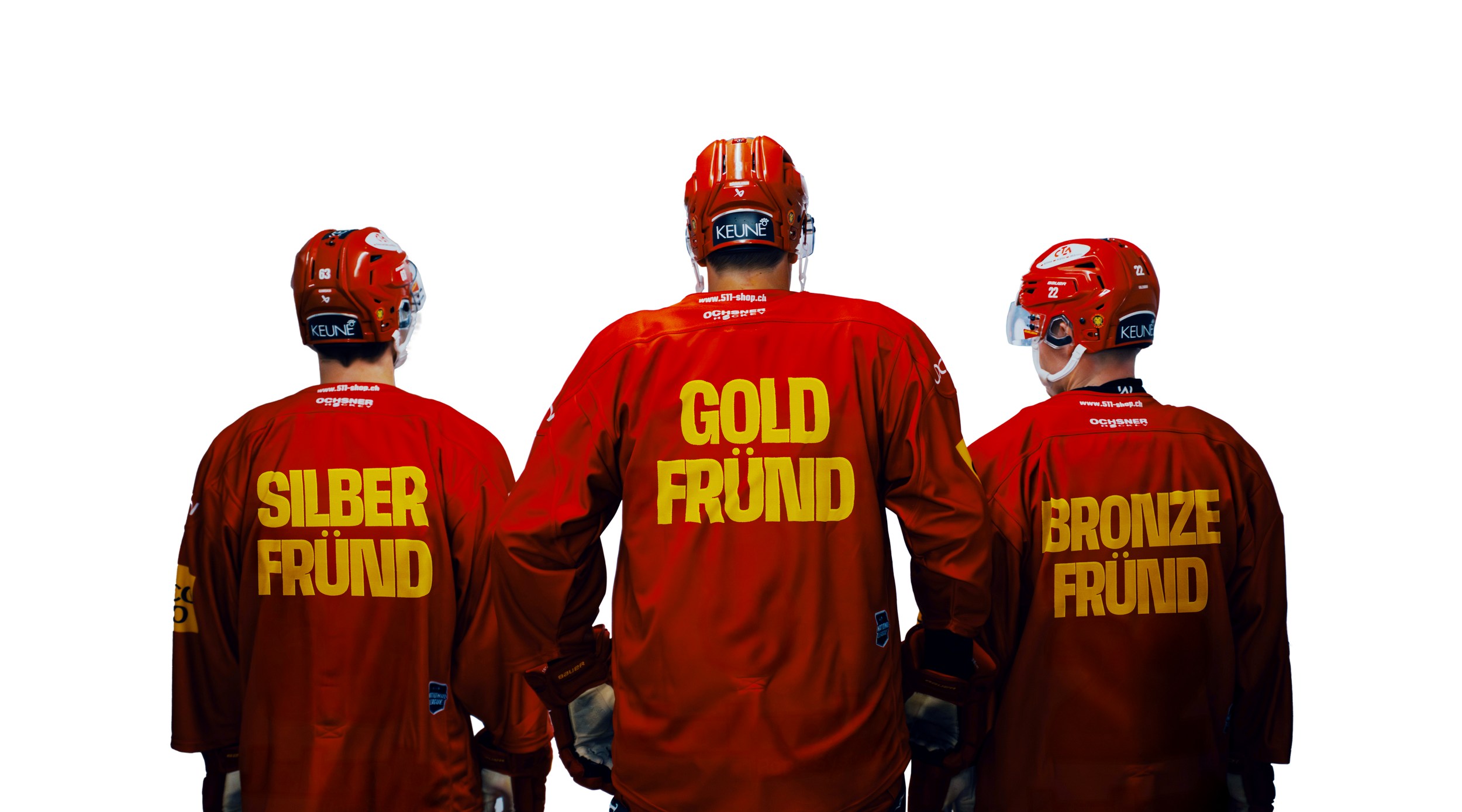Drei Eishockeyspieler mit roten Helmen und roten Jacken, die die Anzeigetafel bei einem Eishockeyspiel anschauen. Die Jacken tragen die Beschriftungen 'Silber', 'Gold', 'Bronze' und 'Freund'.