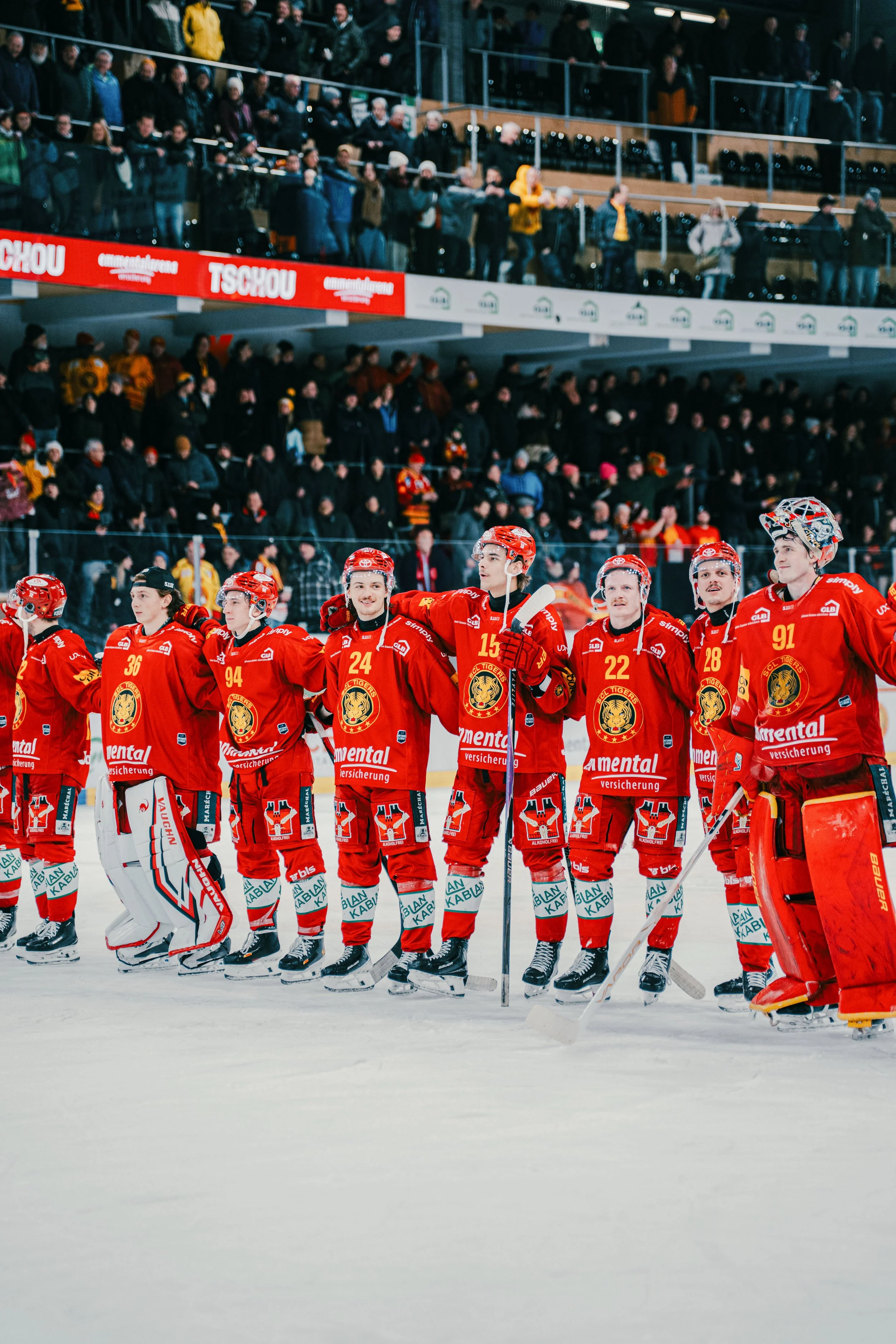 Eishockeyspieler stehen in einer Reihe auf dem Eis, trugen rote Trikots mit einem Löwenlogo, im Hintergrund stehen Zuschauer in der Arena.