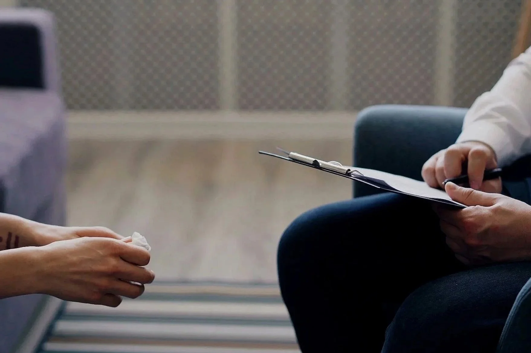 A person holding tissue in hands and another person in a white shirt holding a clipboard with a pen, possibly during an interview or consultation.
