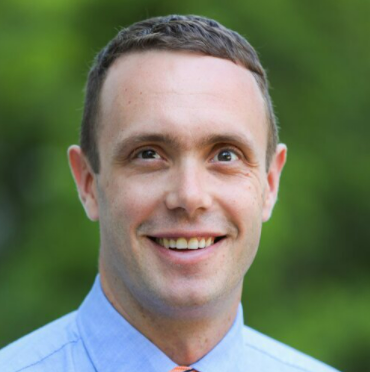 A smiling man with short brown hair wearing a blue collared shirt outdoors with a green background. Member of the AI Aptitude Institute Advisory Board and expert in enterprise AI strategy