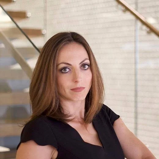 A woman with shoulder-length brown hair, wearing a black top, standing in front of a staircase with wooden steps and a white brick wall. Member of the AI Aptitude Institute Advisory Board and expert in enterprise AI strategy