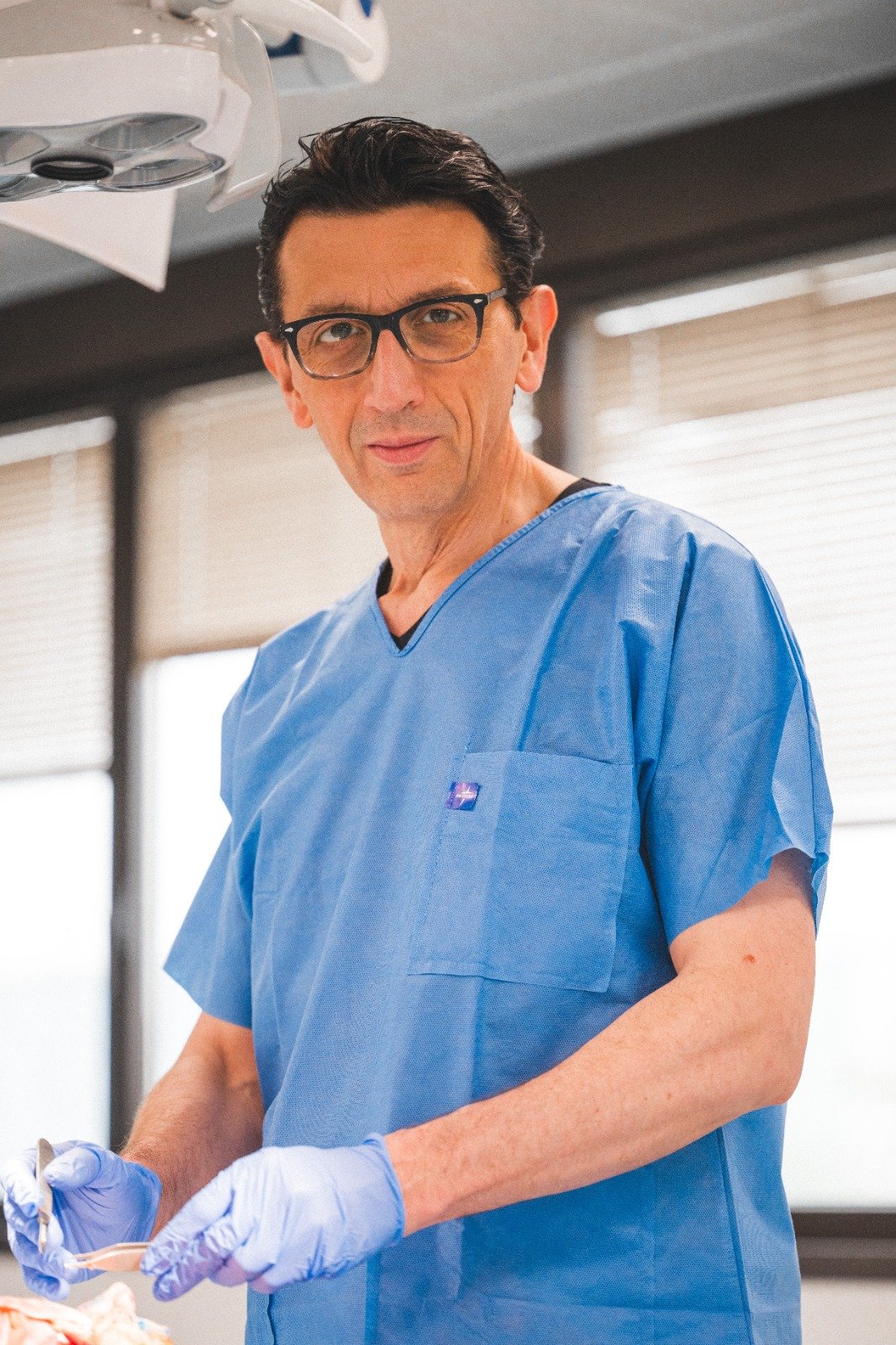 Un uomo in divisa da medico con occhiali, in un ambiente clinico, che tiene uno strumento chirurgico.