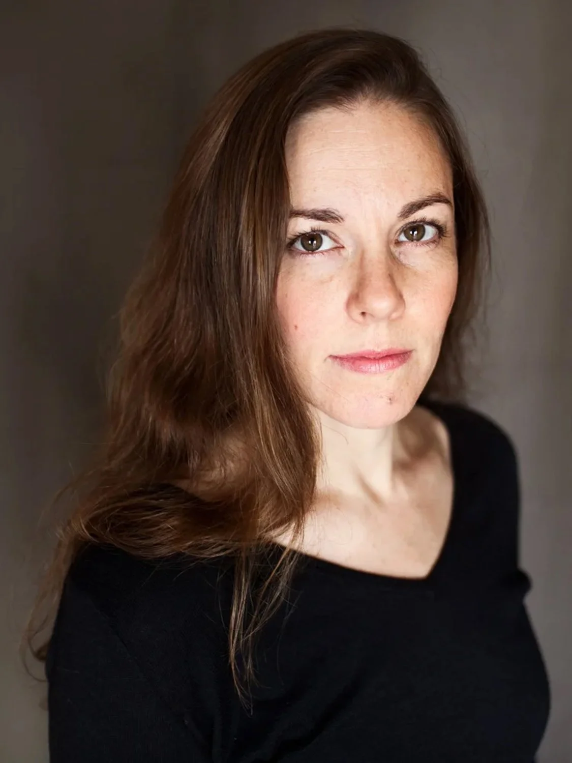 A woman with brown hair and green eyes, wearing a black top, against a dark background.
