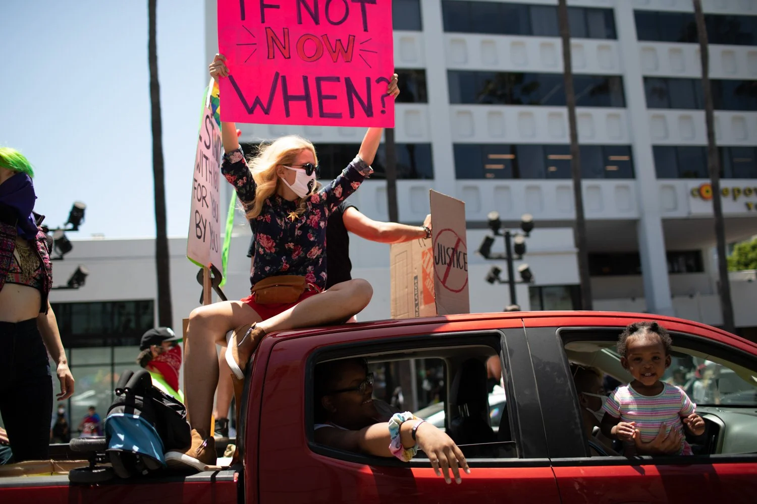Protesters riding in the back of a red truck during a demonstration on a city street, with people holding signs, including one that says 'IF NOT NOW WHEN?', and a large modern building in the background.