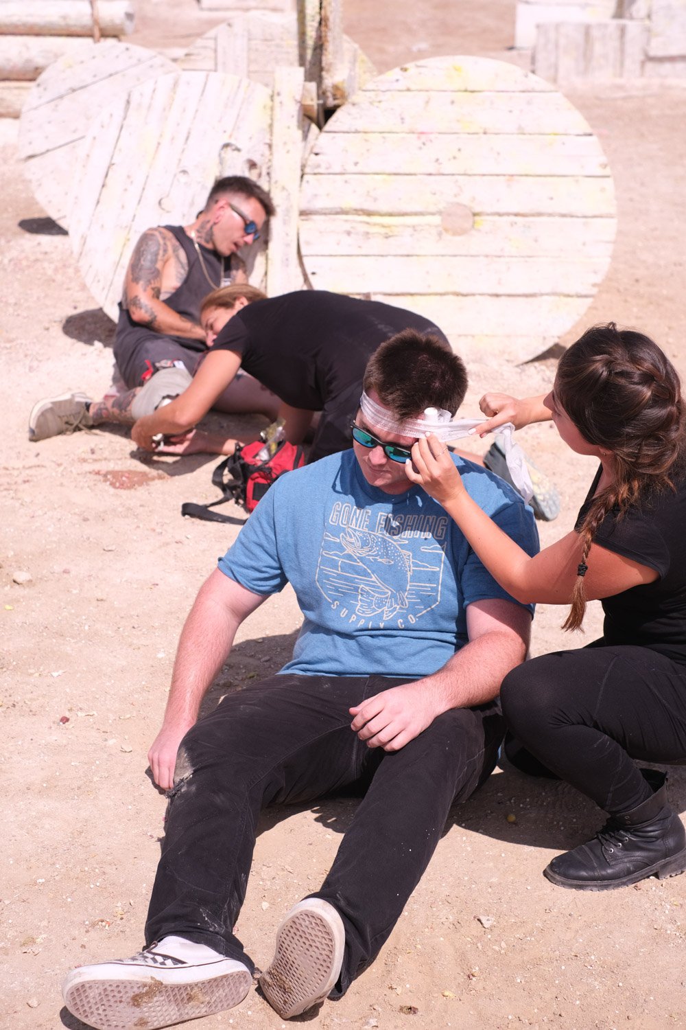A group of young people sitting and lying on the sandy ground with large wooden spools in the background. One person with bandages on his head is sitting while another is leaning over him. One individual appears to be tending to another's injured head, possibly offering first aid or comfort.