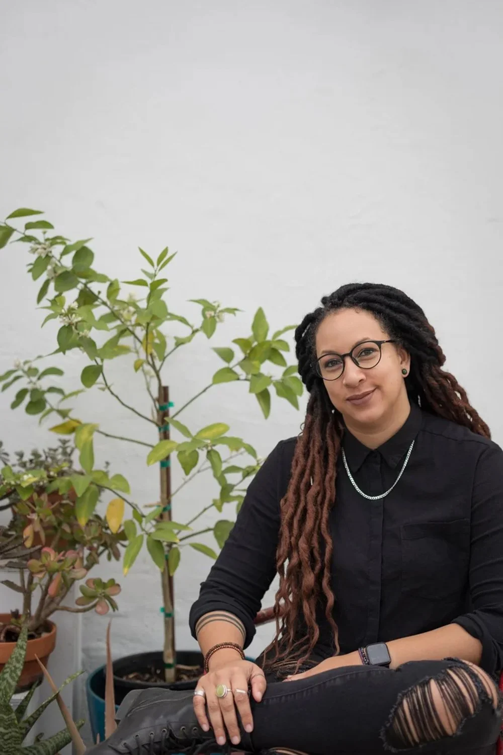 A woman with glasses, dreadlocks, and a black shirt sitting next to potted plants against a white wall.