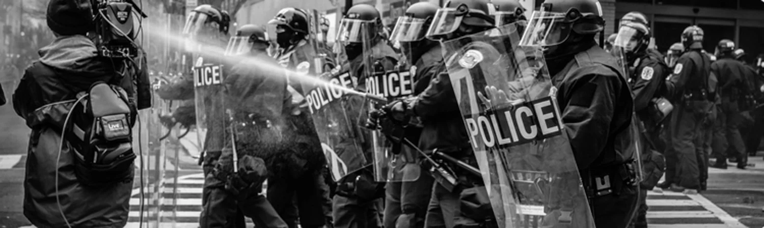 Line of police officers in riot gear holding shields with 'POLICE' written on them, standing in formation on a city street.