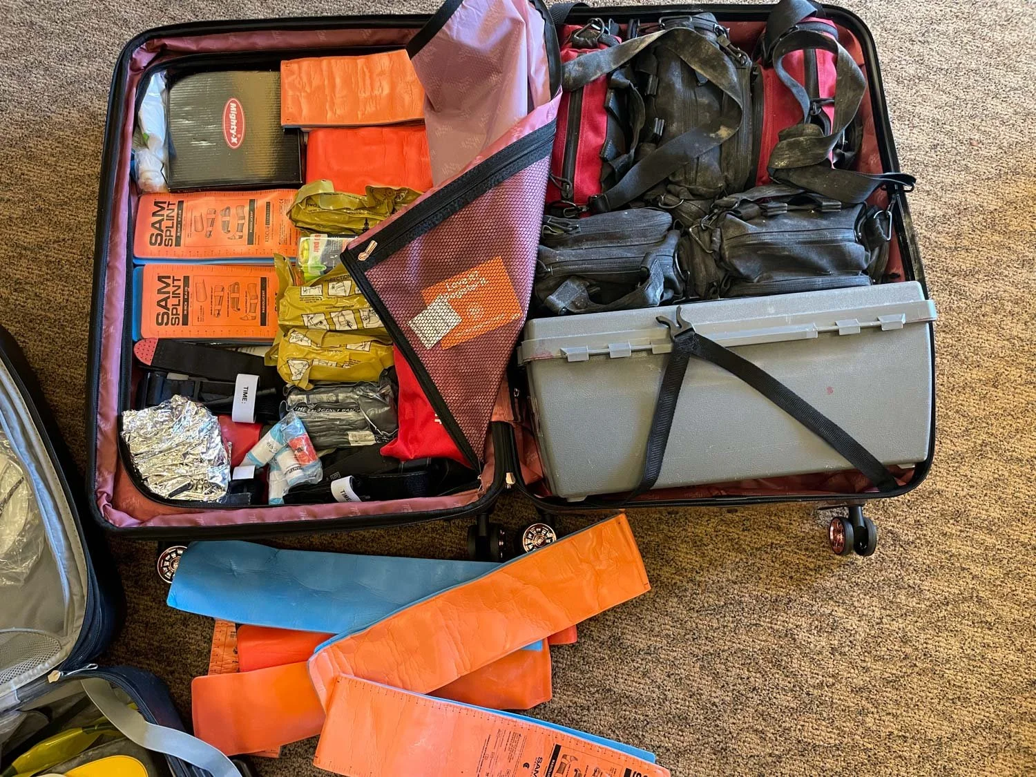 Open suitcase packed with various outdoor and camping gear including a first aid kit, flashlights, rain ponchos, and other supplies, on a carpeted floor.