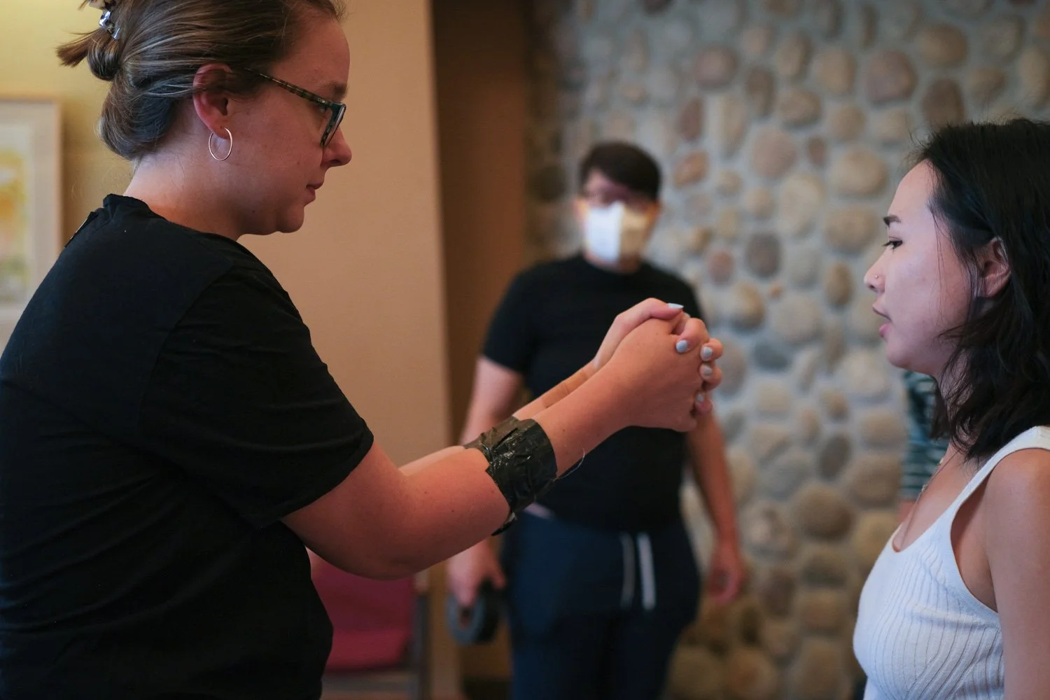 A woman with glasses and hoop earrings holds hands in prayer with a woman in a white tank top, beside a stone wall, while a person in a black shirt and mask stands in the background.