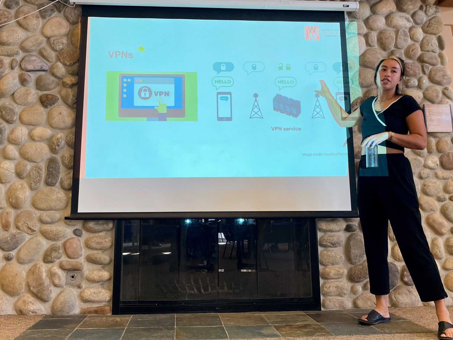 A woman wearing black pants, a black crop top, and sandals stands next to a large screen, pointing at a presentation slide about VPNs. She holds a glass of water, and the background features a decorative stone wall.