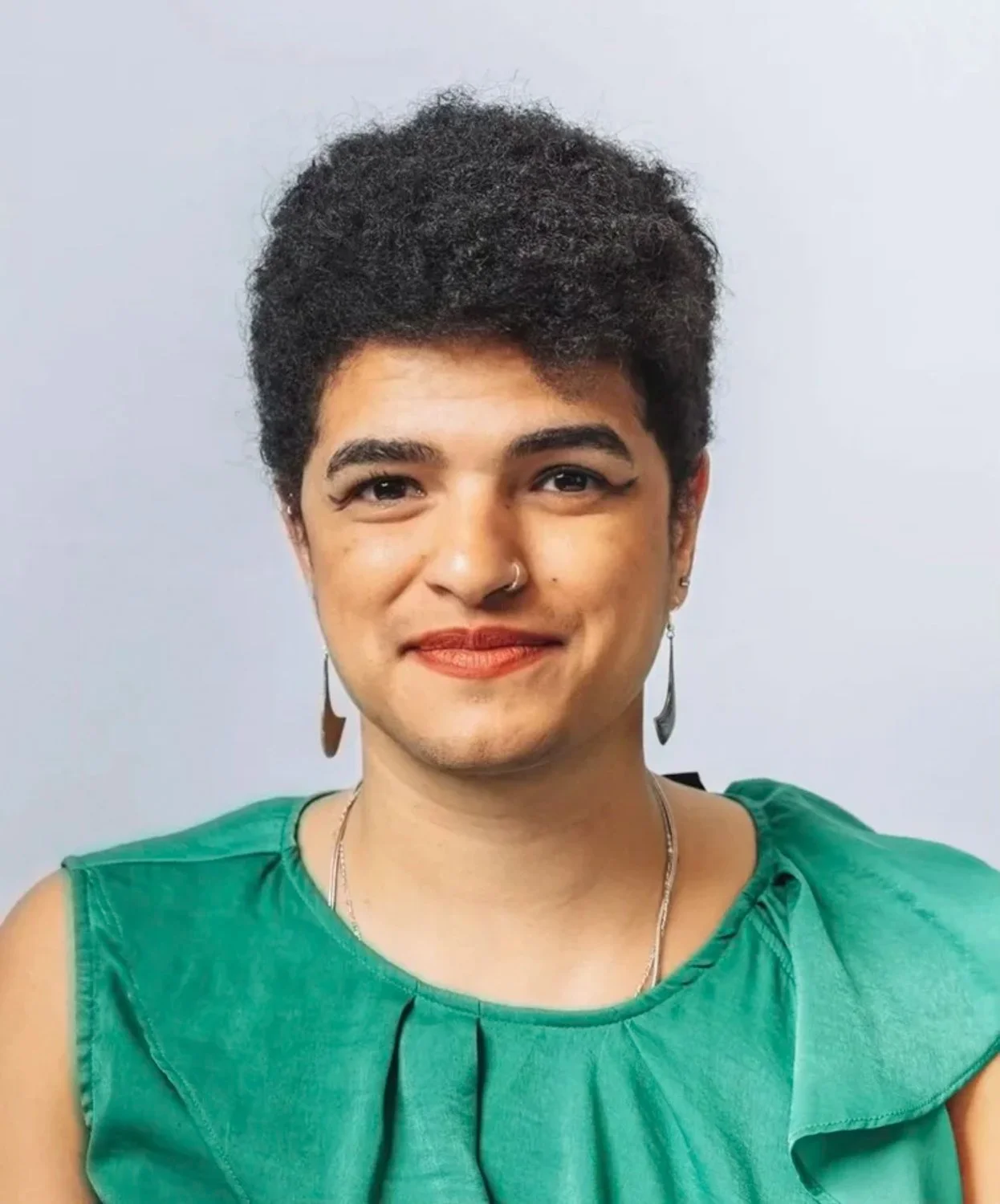 A person with short curly dark hair, wearing a green sleeveless top, silver jewelry, a nose ring, and earrings, smiling at the camera against a neutral background.