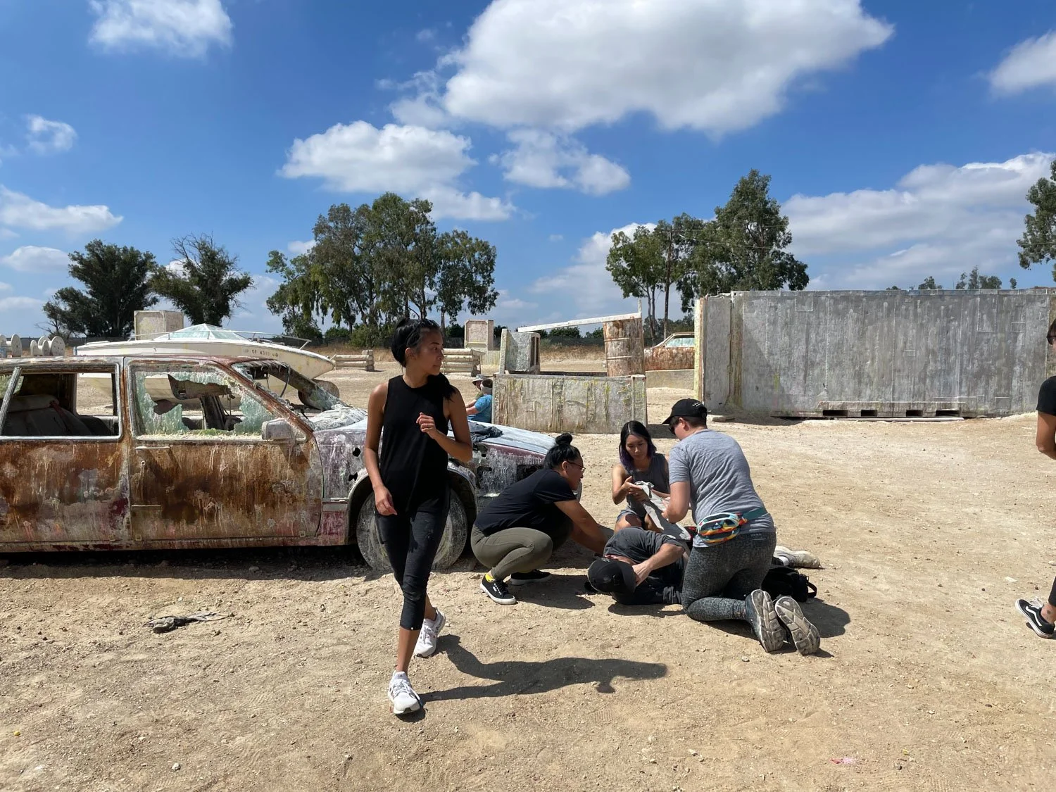 Group of five people, four women and one man, gathered around a person lying on the ground, possibly providing first aid or assistance, beside a rusty, abandoned car in an outdoor setting with a clear blue sky and trees in the background.