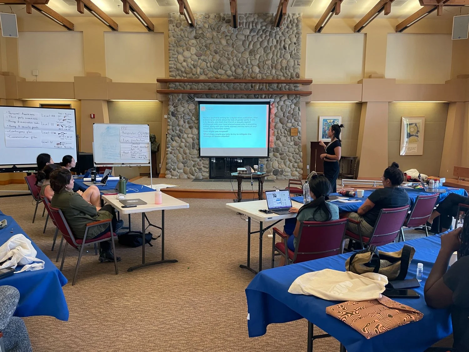 A woman presenting at a conference with an audience seated at tables. The room features a stone fireplace, wooden beams, and large screens displaying presentation slides. Attendees have laptops, notebooks, and water bottles.