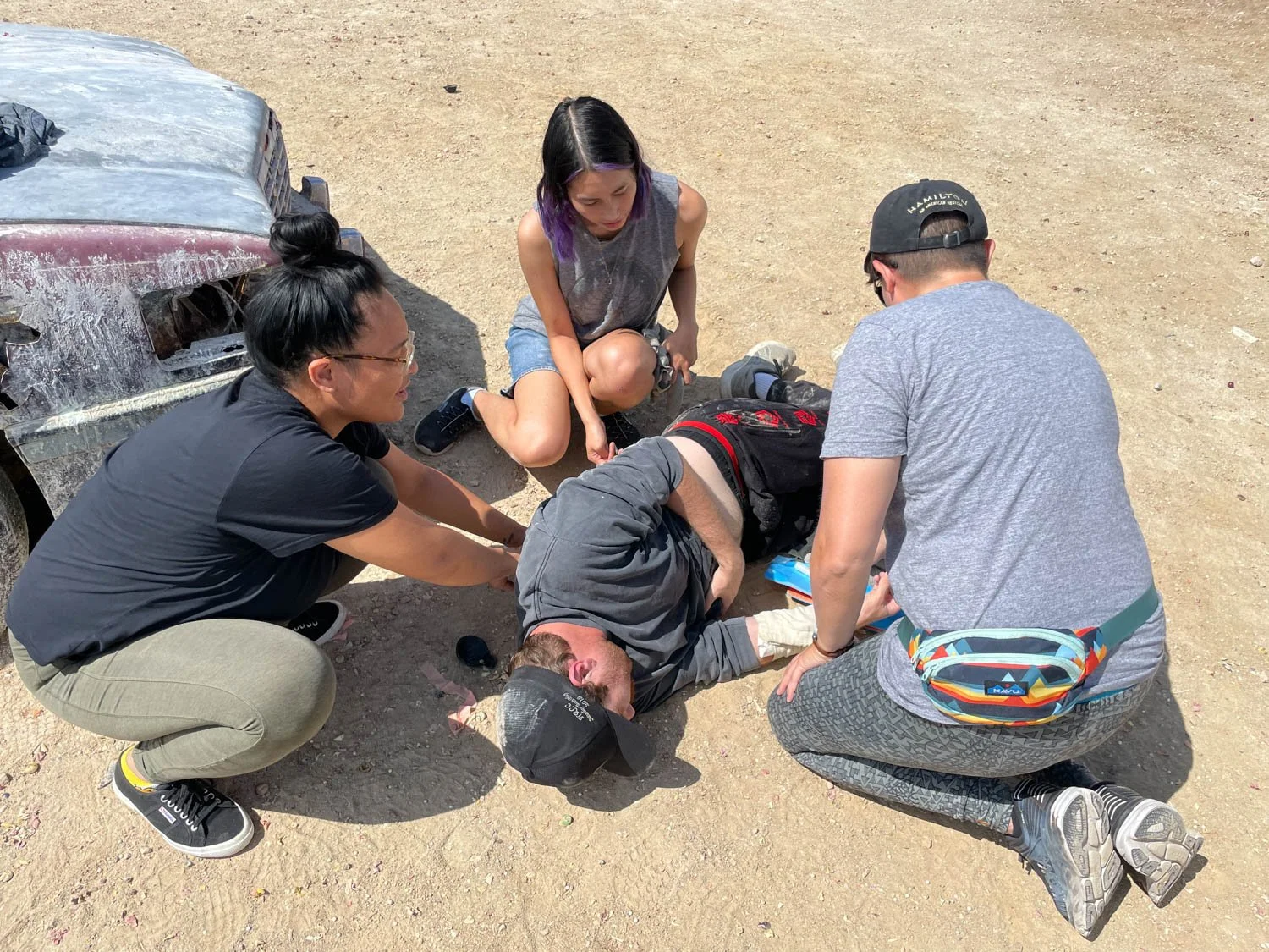 A group of five people attending to an injured man lying on the ground in a desert area. They are near a damaged vehicle, with three individuals crouching and one girl sitting on her knees, all focused on helping the man.