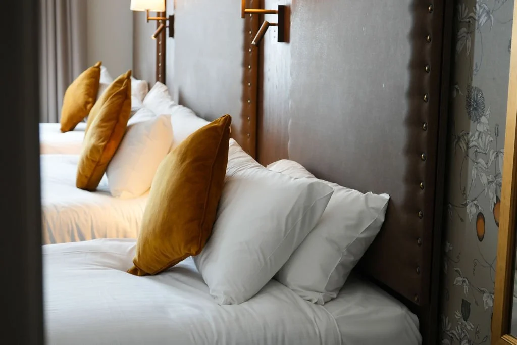 A neatly made bed with white pillows and white linens, viewed through a small opening or door, with a dark wood headboard and a wall-mounted pink and beige striped wall lamp.
