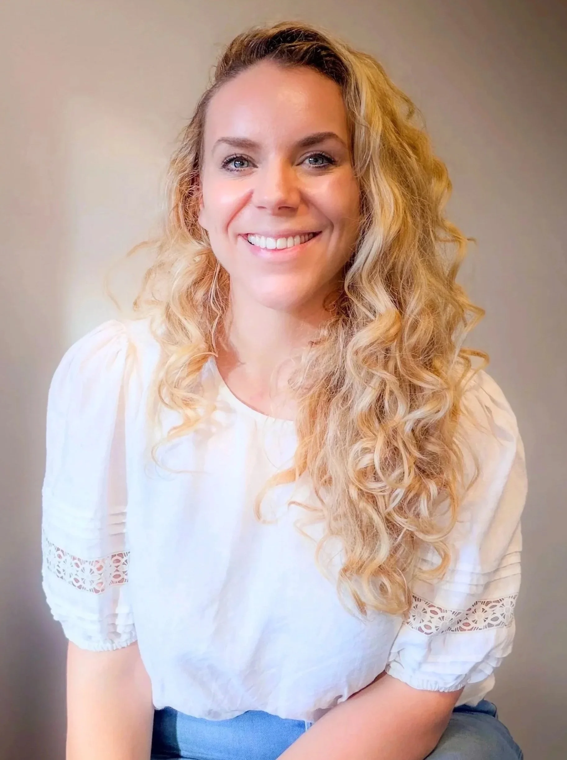 A woman with long, curly blonde hair, blue eyes, and a bright smile, wearing a white lace-sleeve blouse, sitting against a plain background.