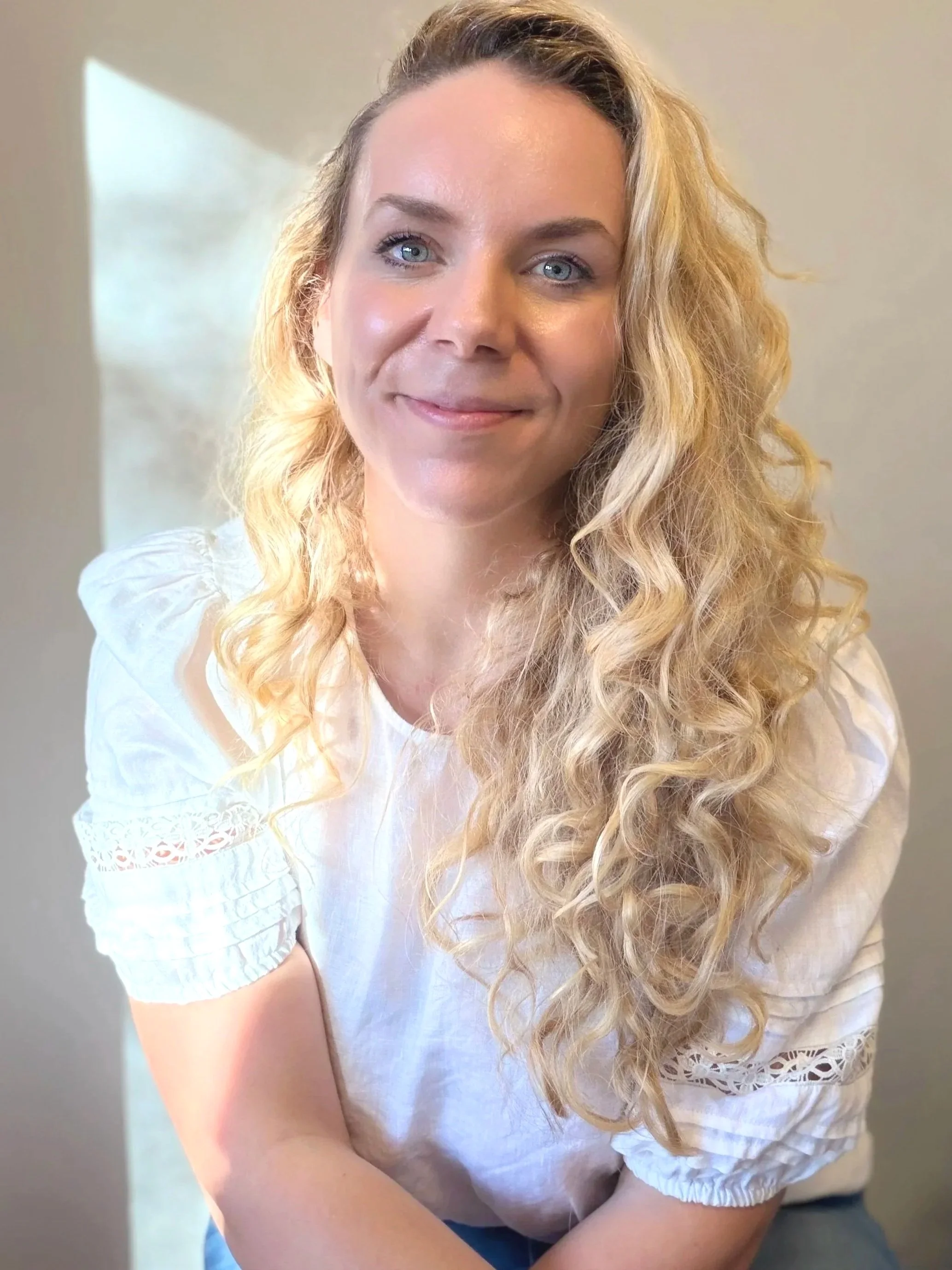 A woman with long curly blonde hair, blue eyes, and fair skin, wearing a white top with embroidered details on the sleeves, smiling at the camera.