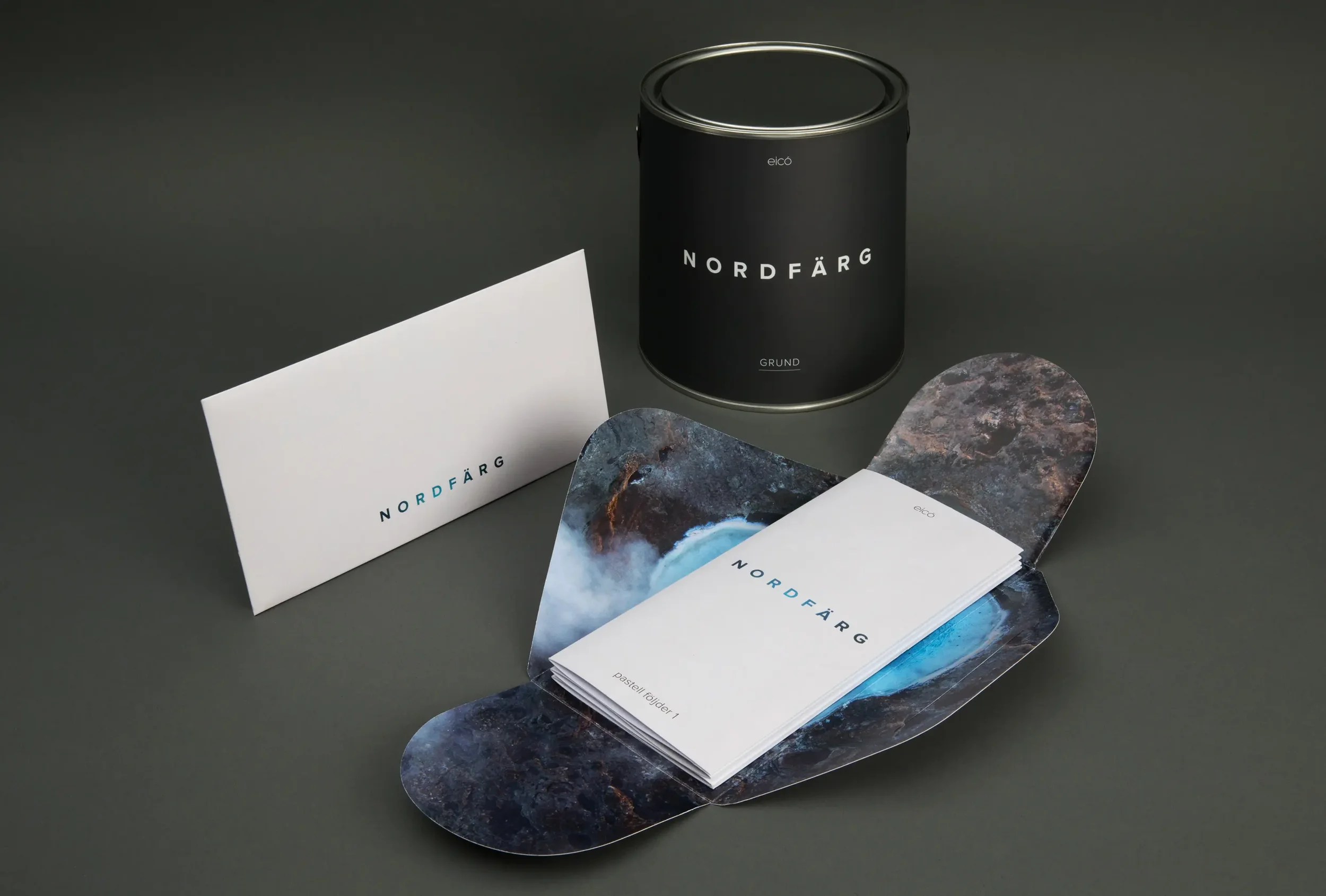 A black cylindrical paint can labeled 'NORDFÄRG', with a white rectangular sample card beside it displaying 'NORDFÄRG' and a colorful abstract landscape design inside a curved portion of the card, all on a dark gray background.