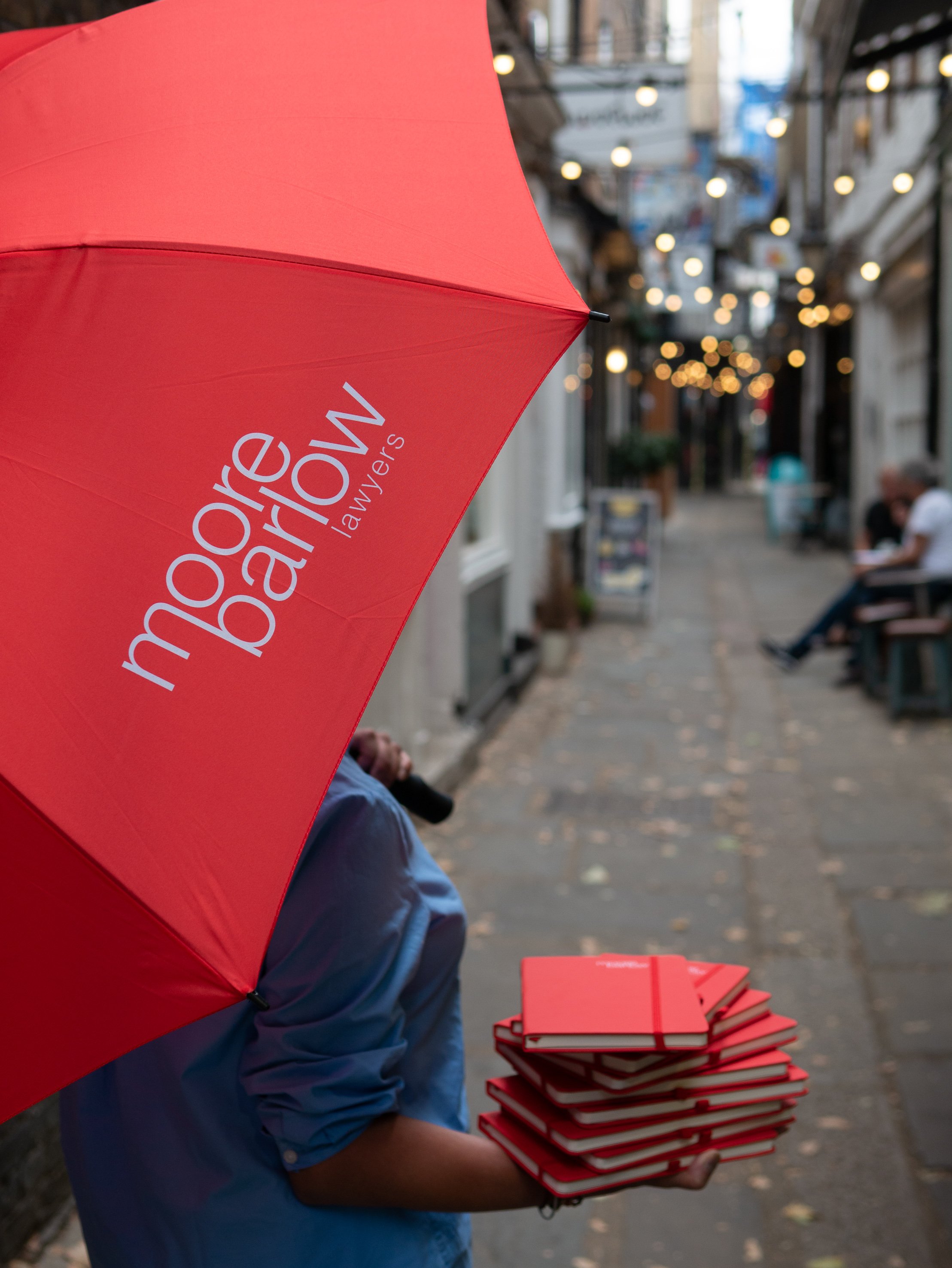 Moore Barlow merchandise. Person holding a stack of red notebooks and an open red umbrella with white text, walking on an outdoor street at dusk with blurred hanging lights and pedestrians.