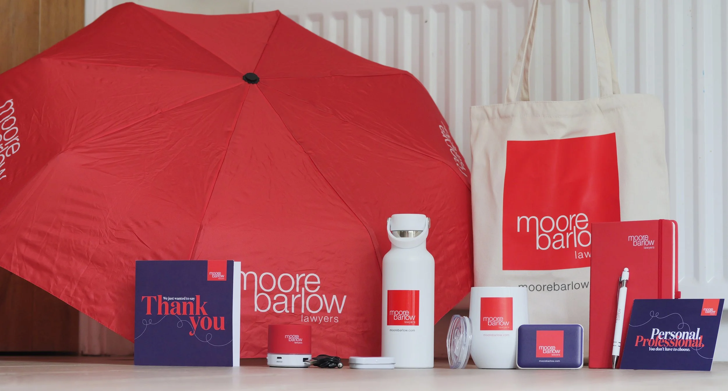 Marketing display for Moore Barlow Lawyers. Items include a red umbrella, a tote bag, a water bottle, card, post card, tote bag, speaker, and various branded promotional materials, all with the company's logo and colours.