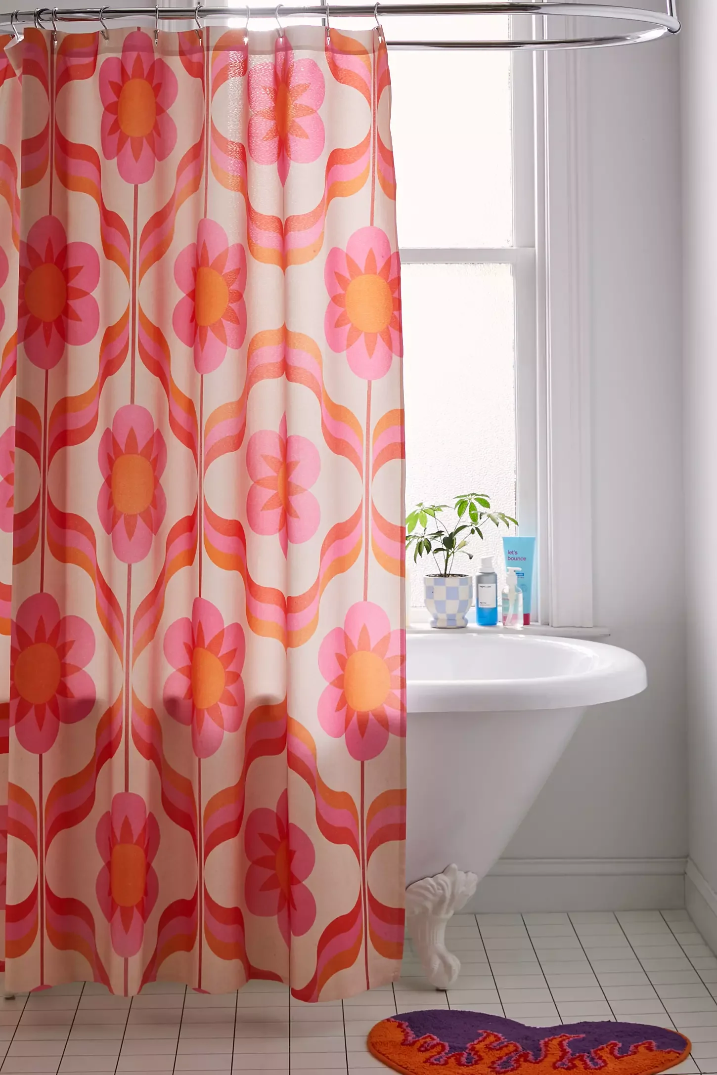 A bathroom with a floral-patterned pink and orange shower curtain, a white clawfoot bathtub, a window with frosted glass, and a small plant on the windowsill. There's a purple and orange rug on the tiled floor.