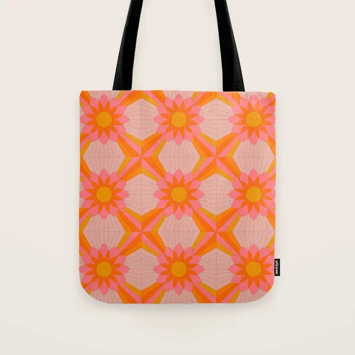 Society6 – Who loves the sun art print. Pink tote bag with a floral geometric pattern featuring pink and yellow flowers and orange accents, hanging by black straps on a white background.
