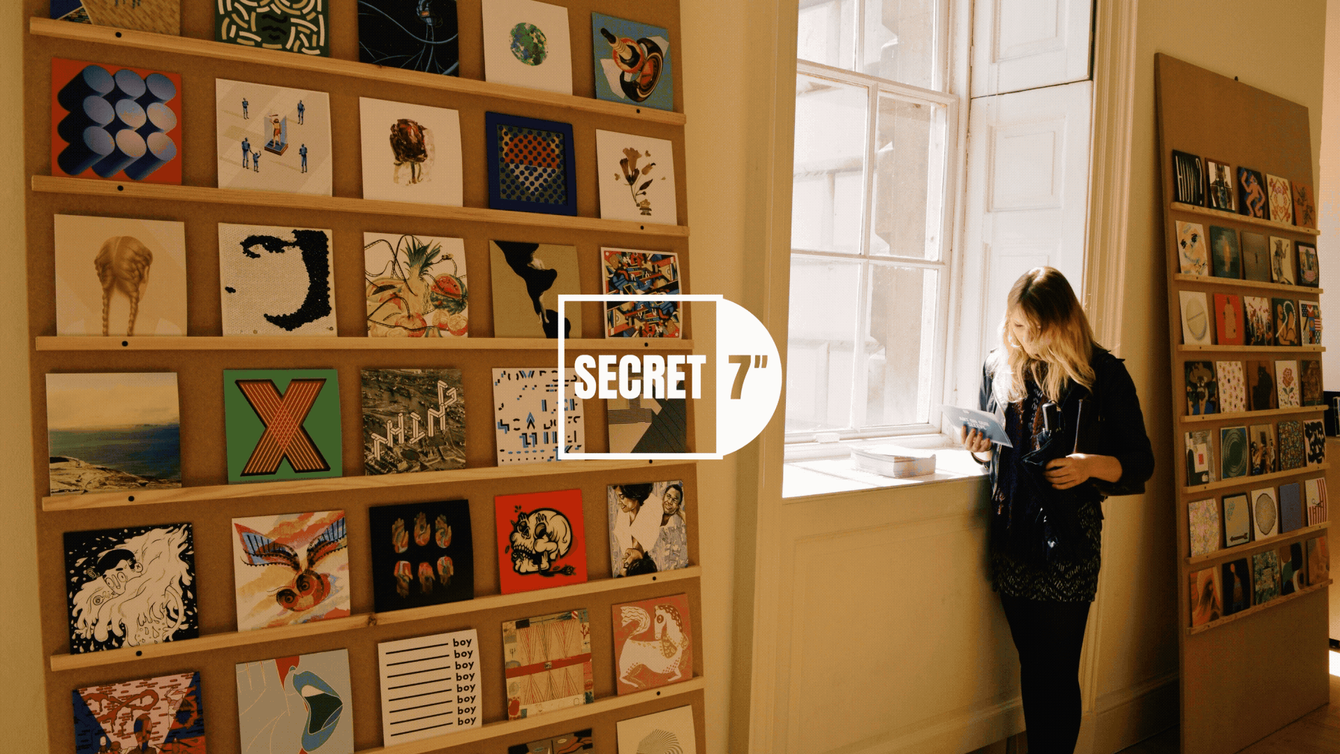 Secret 7" exhibition. A woman in black clothing stands near a window in an art gallery, reading a booklet, with two shelves of colourful vinyl artwork behind her.