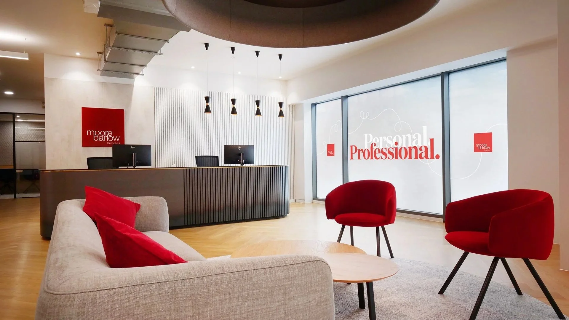 Modern law office reception area with beige sofa and red pillows, red chairs, a reception desk with two computer monitors, and a large window with frosted glass displaying the words "Personal. Professional." and the law firm's logo, Moore Barlow Lawyers.