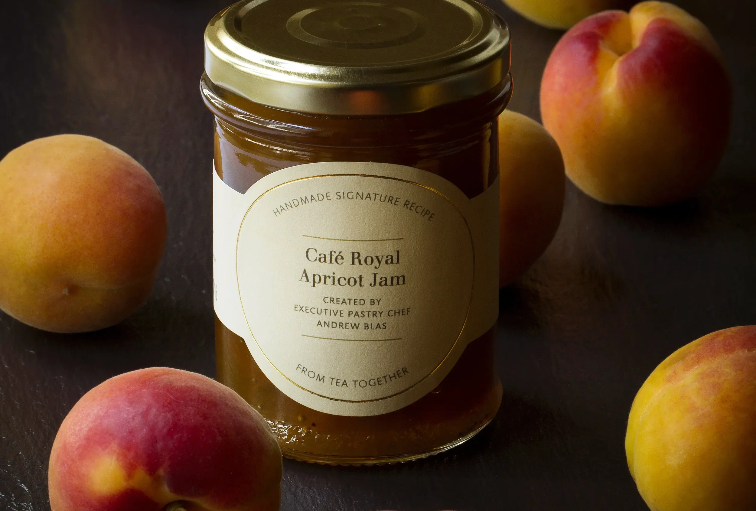A jar of apricot jam with a gold lid, labeled 'Café Royal Apricot Jam,' surrounded by fresh peaches and apricots on a dark surface.
