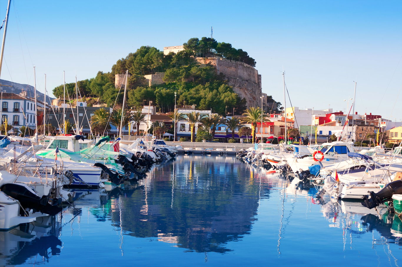Un port de plaisance avec des bateaux amarrés, face à un village avec des maisons colorées et un château sur une colline verte, sous un ciel clair.